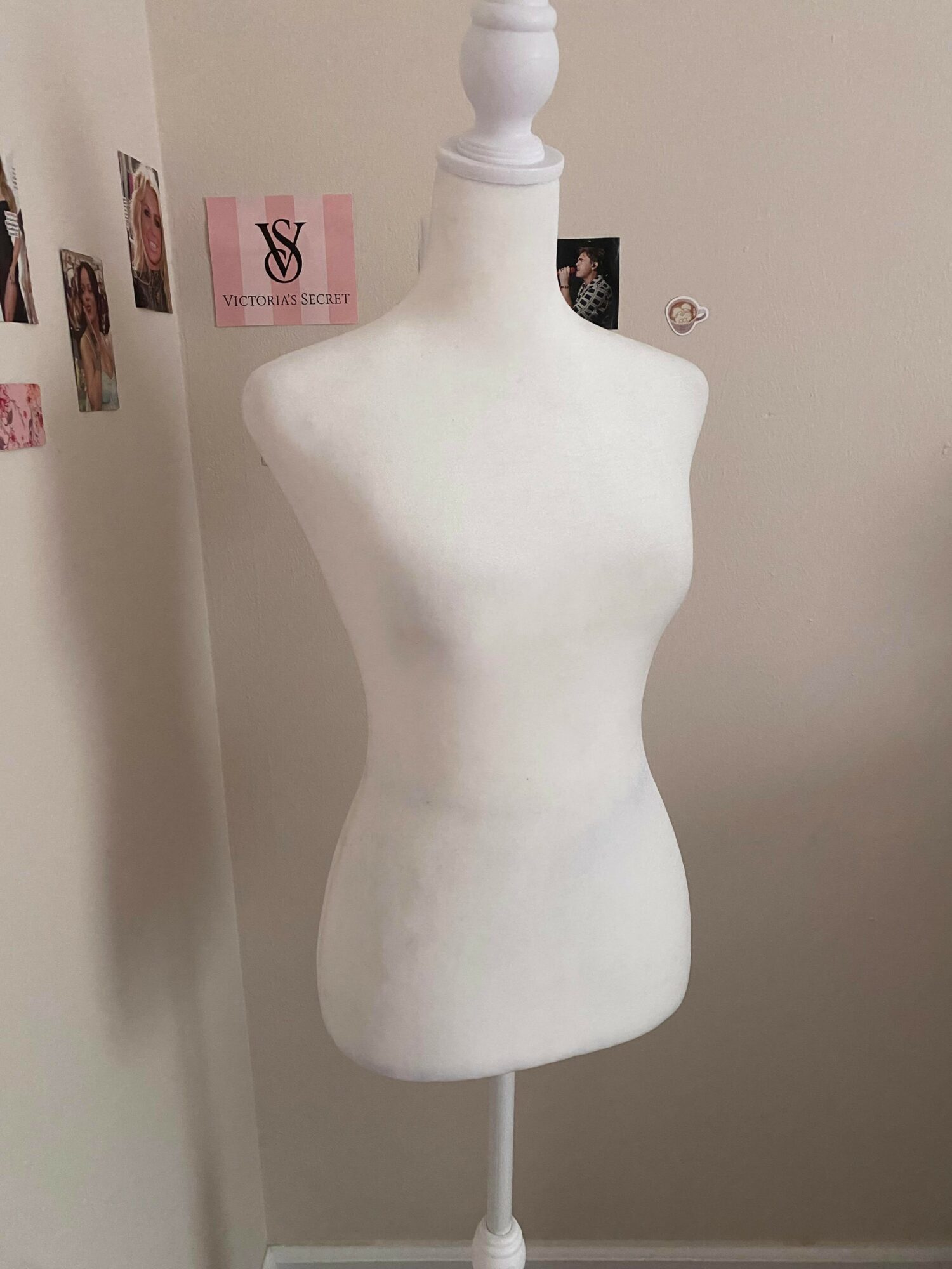 White mannequin torso on stand against a wall with photos and pink paper, no head or arms visible.