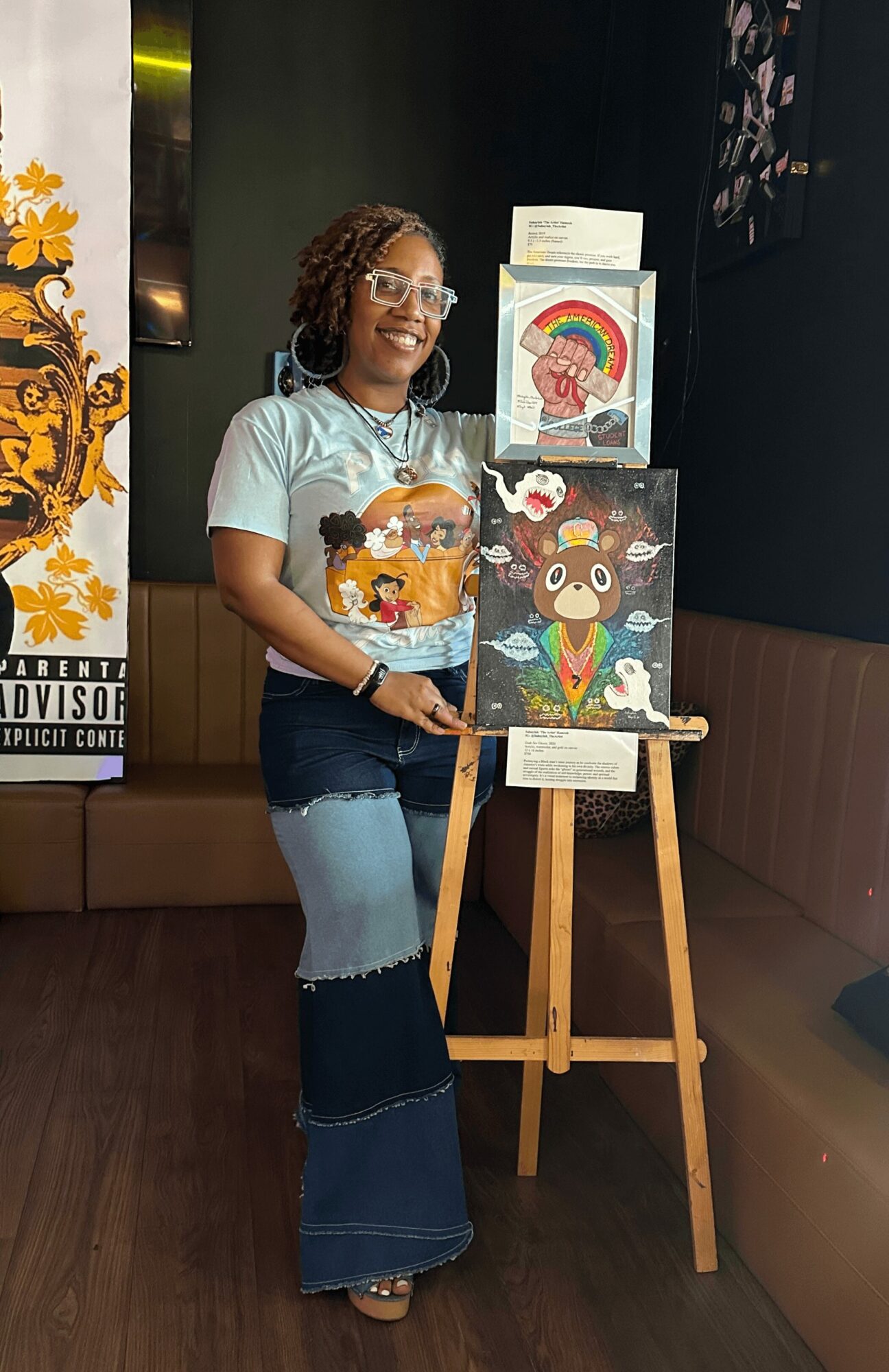 Young girl with glasses and curly hair stands next to colorful artwork on an easel, smiling indoors.