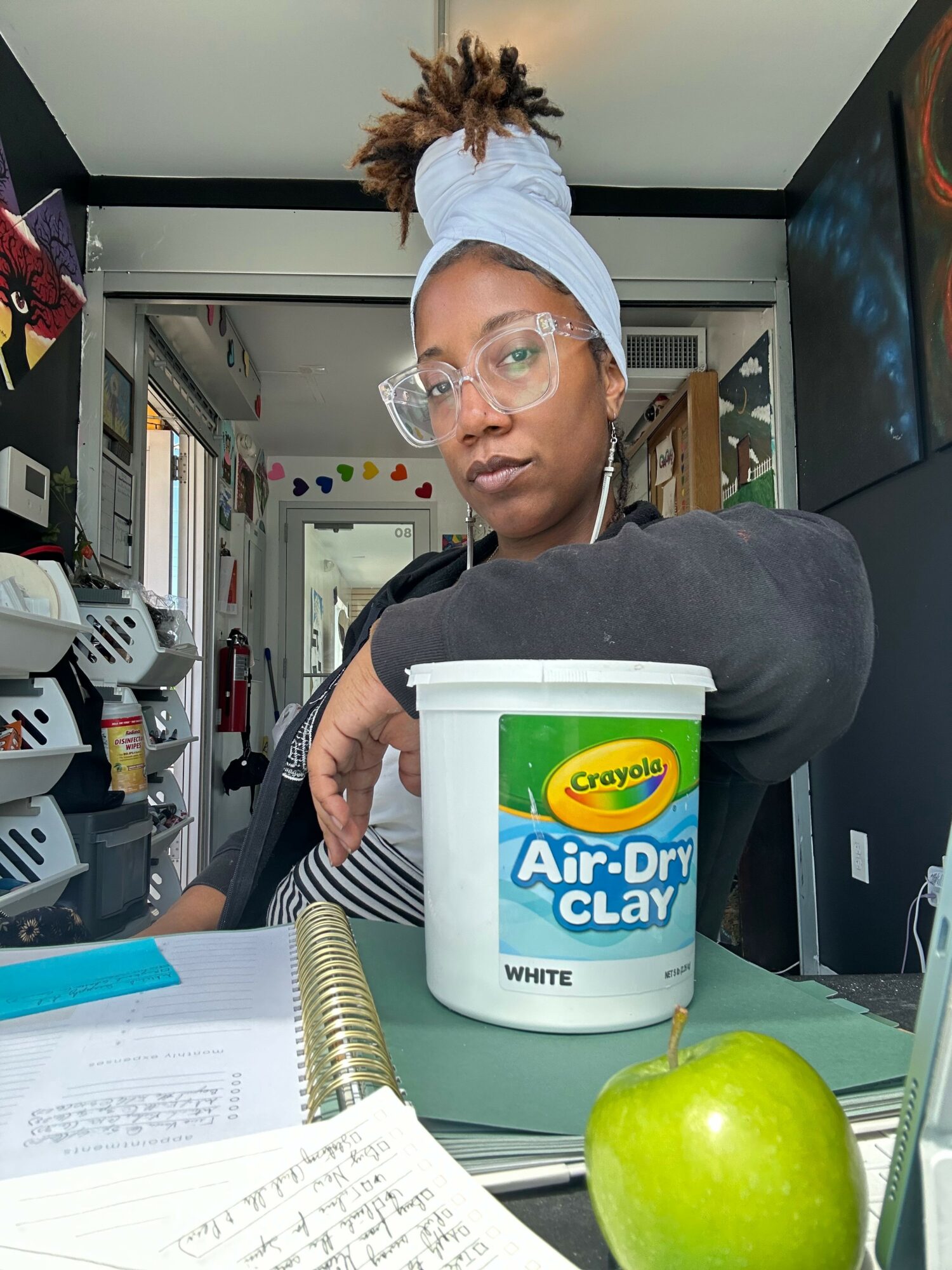 Person with glasses and headscarf in a kitchen holding a container of air-dry clay, with a green apple and notebook on table.