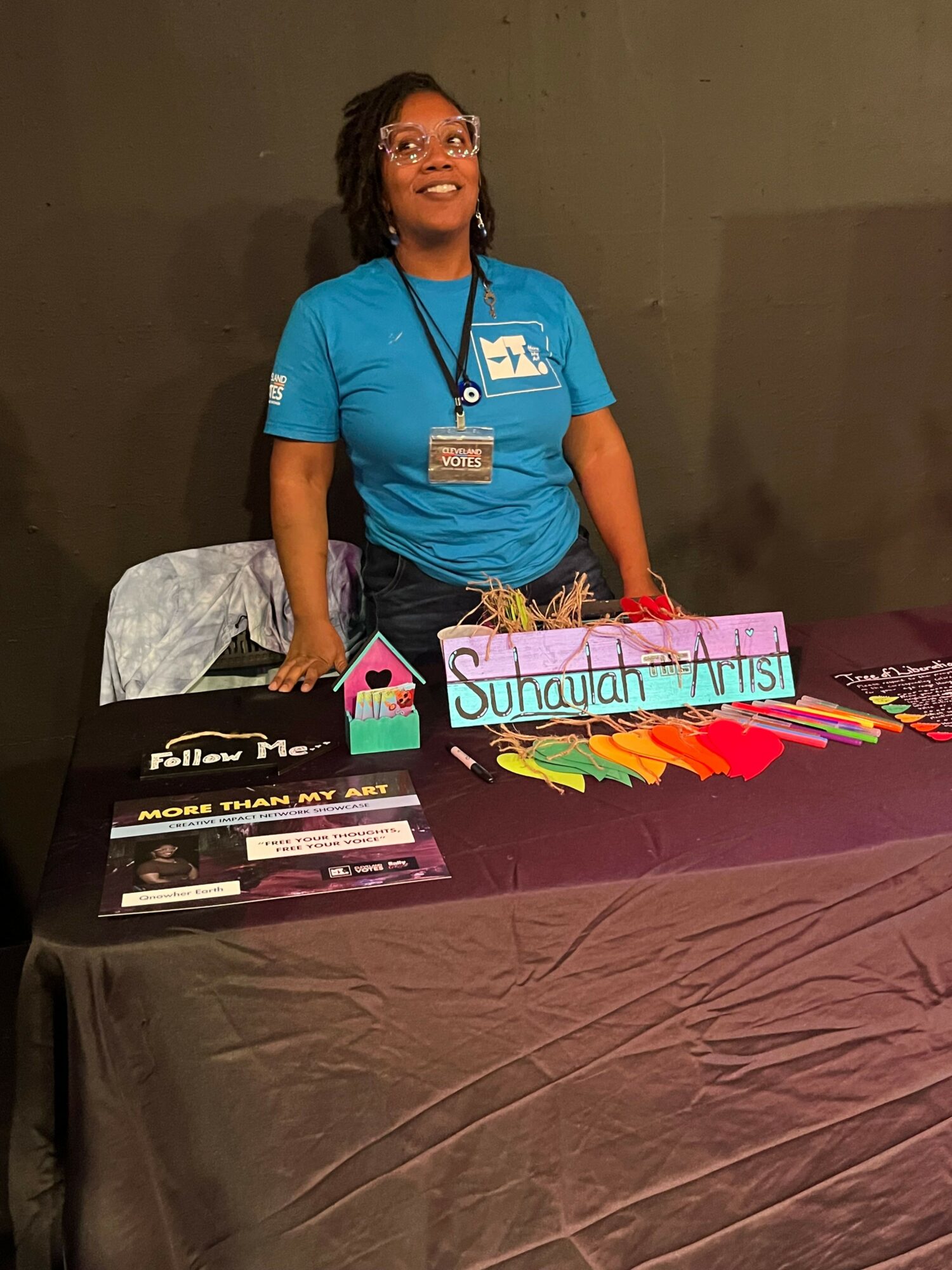 Woman standing next to a table with colorful items and a sign, indoors with a dark background.