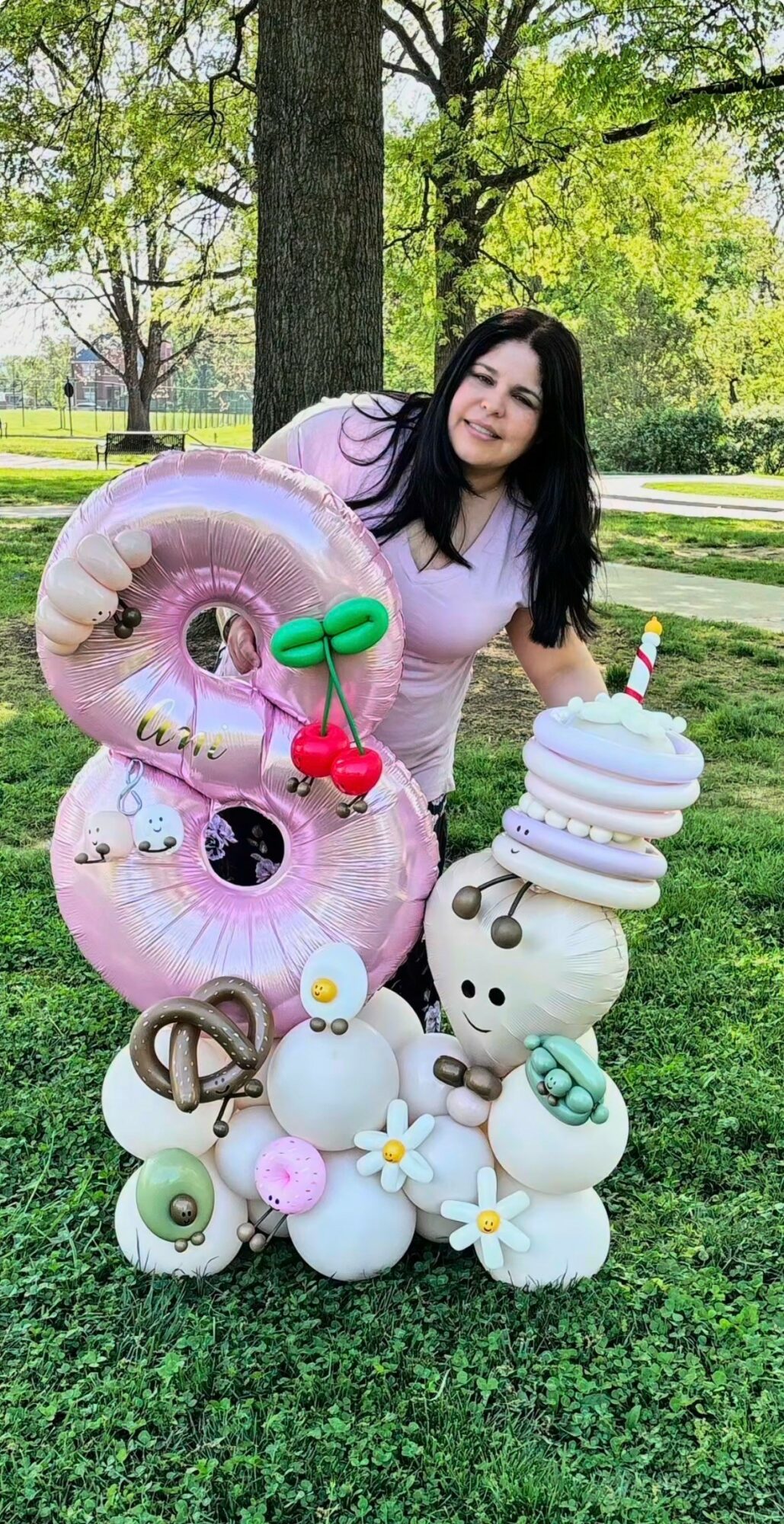 Young woman outdoors with balloon animals and decorations, standing on grass near trees, smiling, sunny day.