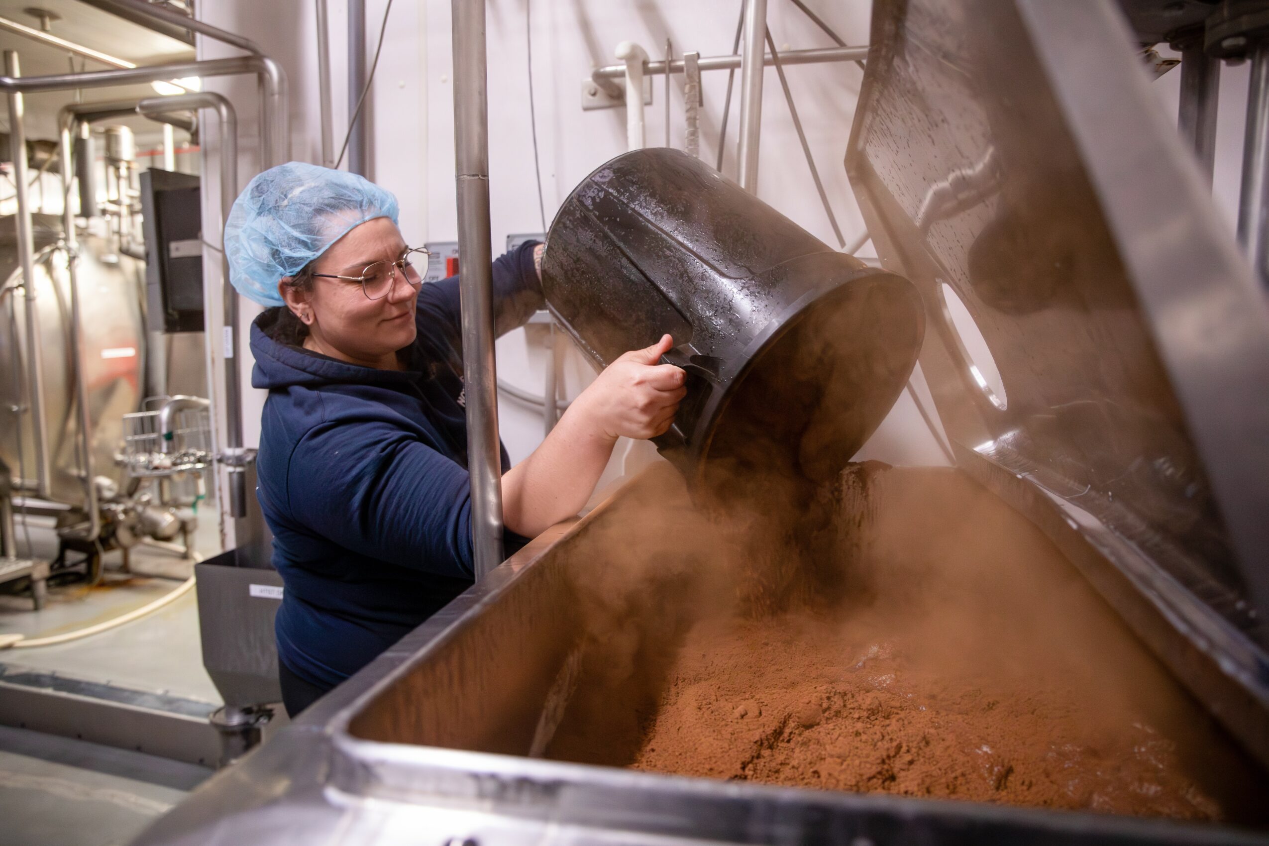 Person wearing a hairnet and glasses pours a dark powder into a large industrial container, with steam or dust rising.