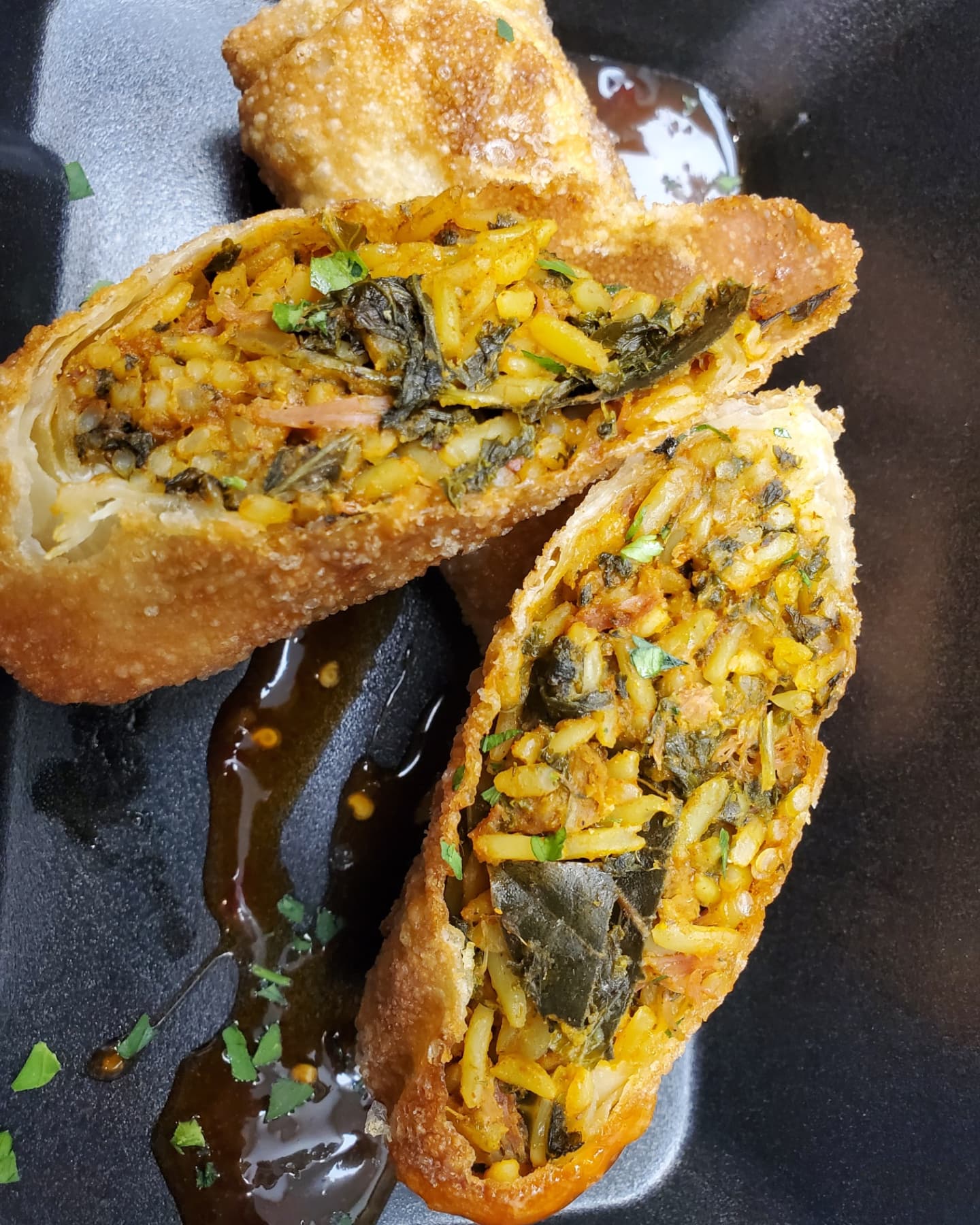 Two fried stuffed pastries with yellow filling and green herbs inside, on a black plate with sauce and a fried item in the background.