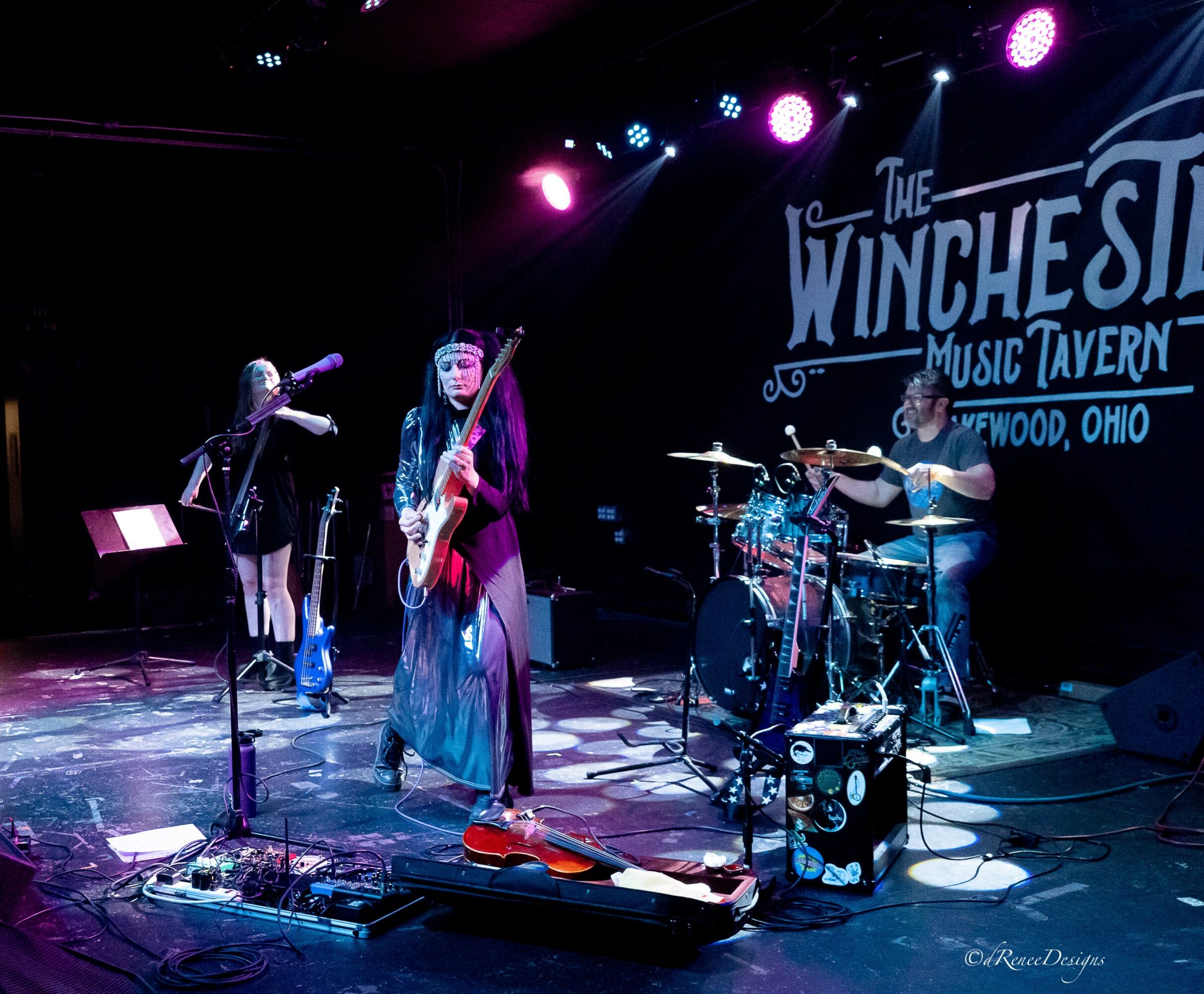 Three musicians perform on stage at The Winchester Music Tavern in Ohio, with instruments and stage lighting visible.