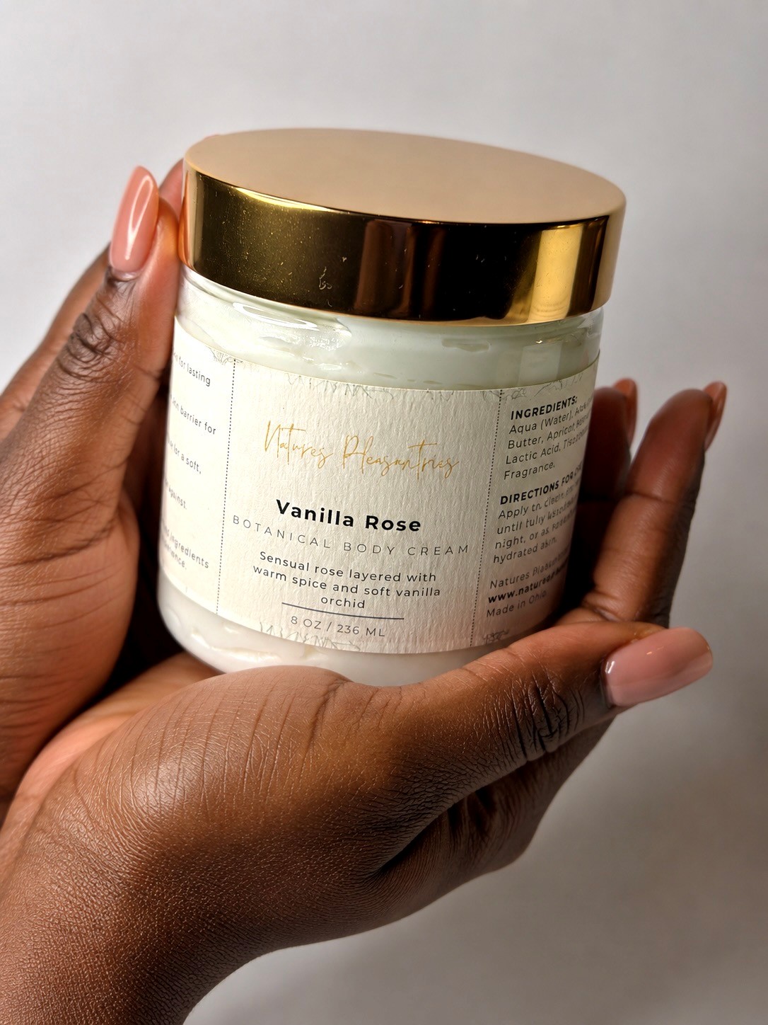 Hand holding a jar of vanilla rose scented body cream with a gold lid, against a plain background.