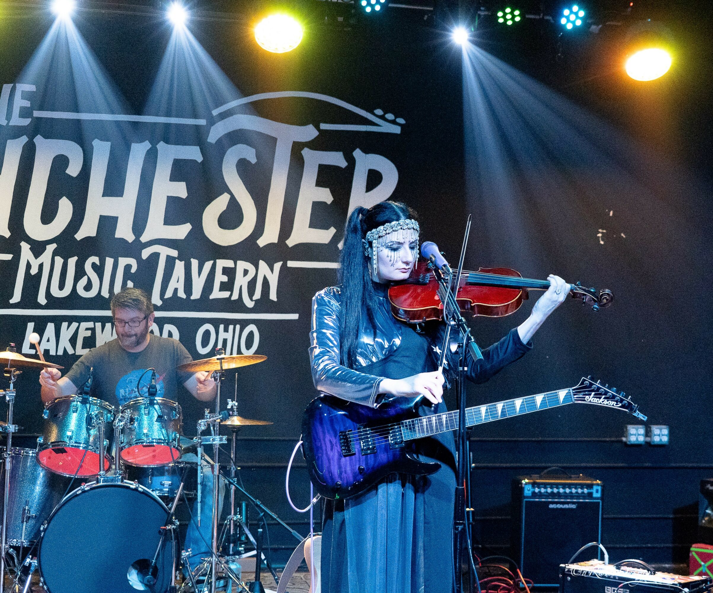 Female musician playing violin and guitar on stage with drummer in background, colorful stage lights, and a sign reading 'The Winchester Music Tavern'.