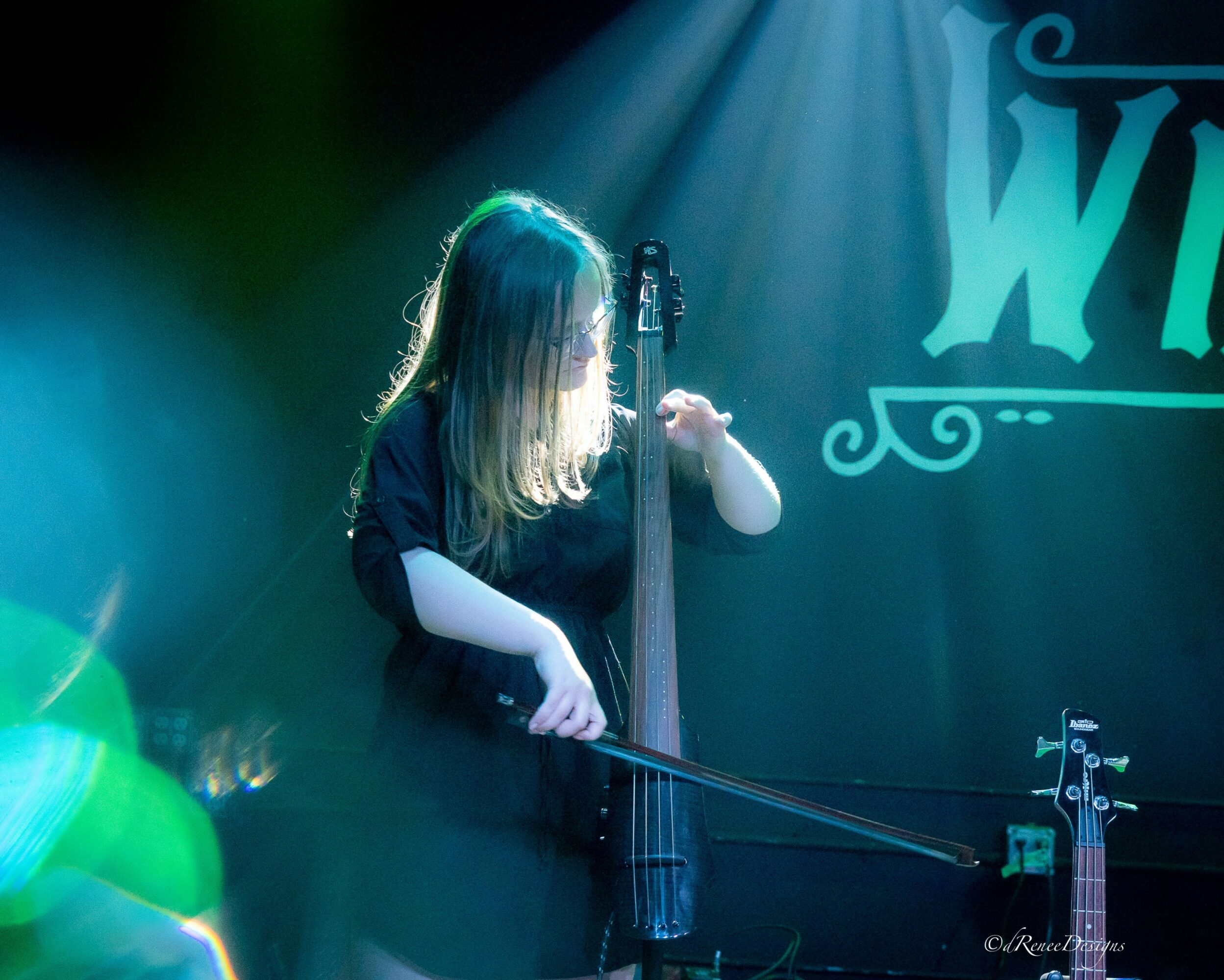 Person playing a string instrument on stage with blue and green lighting, and a partially visible sign in the background.