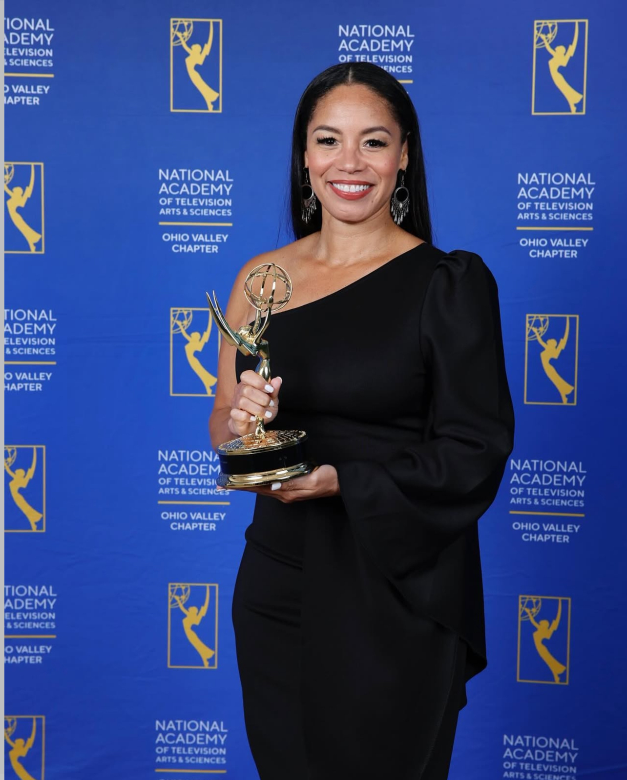 Woman smiling holding an award trophy in front of a blue backdrop with logos and text.