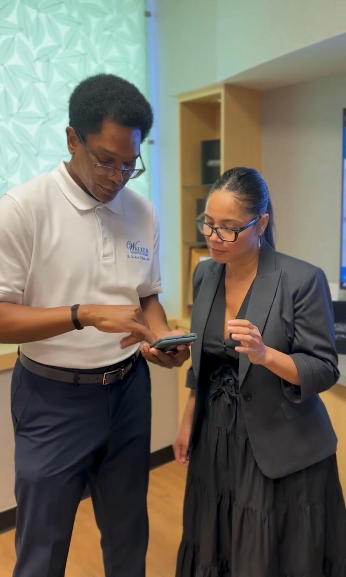 Two people, a man and a woman, stand indoors looking at a smartphone together, both wearing glasses and formal attire.