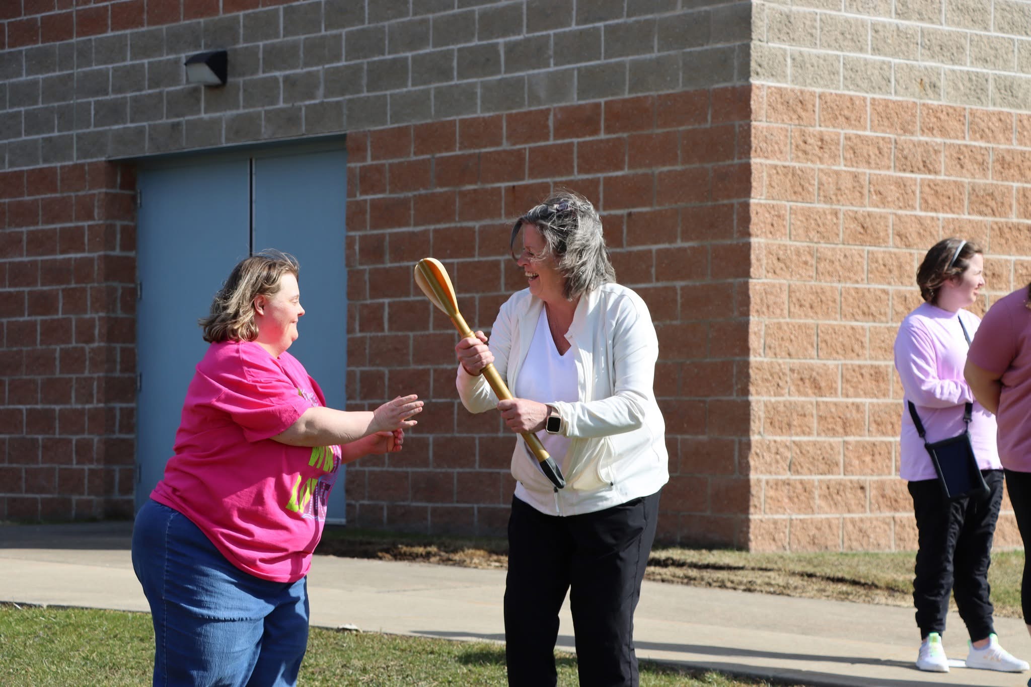 Two women exchanging a wooden spoon outdoors near a brick building, with two other women in the background.