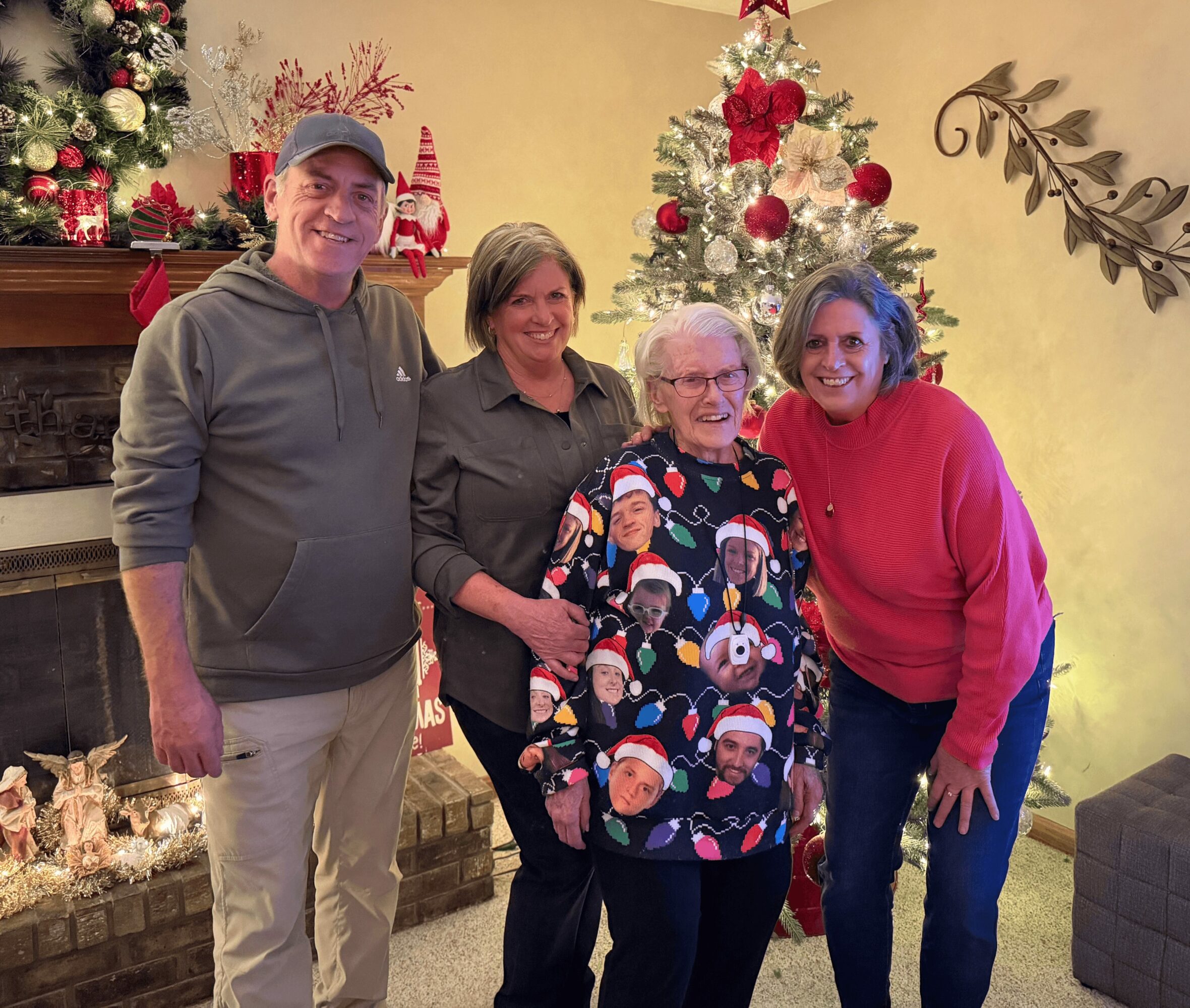 Four people standing together in front of Christmas trees and decorations, smiling indoors during holiday celebration.