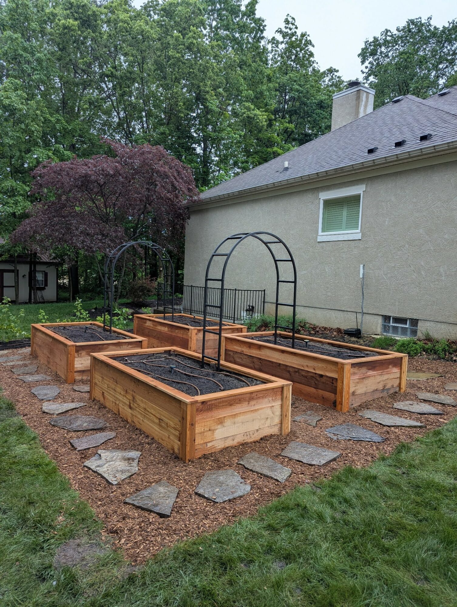 Garden with five raised wooden beds, stone pathway, arch trellis, and house in background, surrounded by trees.