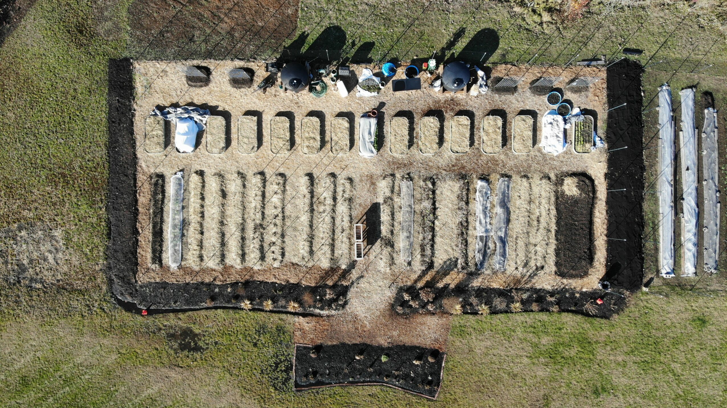 Aerial view of a rectangular structure with rows of objects inside, surrounded by grass and a dirt path, with additional structures nearby.
