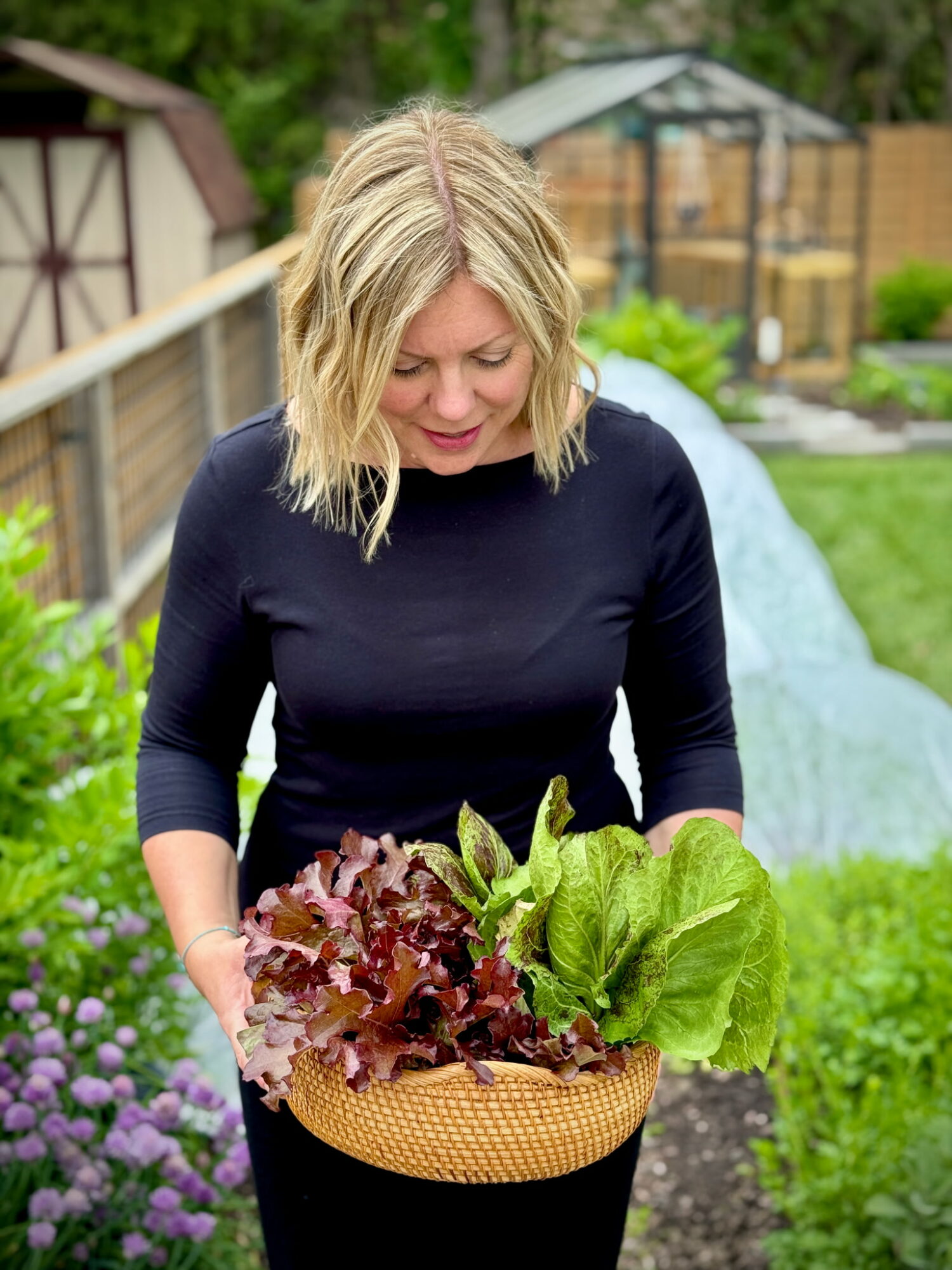 Woman with blonde hair holding a basket of leafy greens and lettuce in a garden.