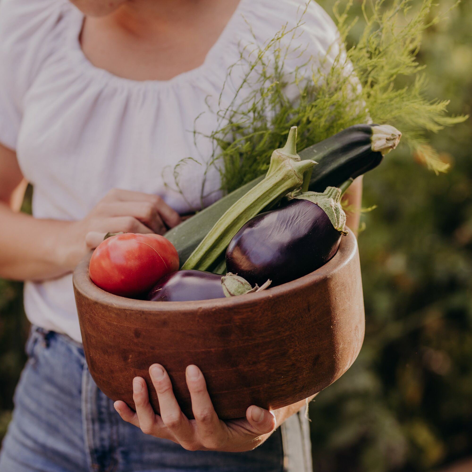 Person holding a wooden bowl filled with vegetables including eggplants, an apple, and leafy greens, outdoors.