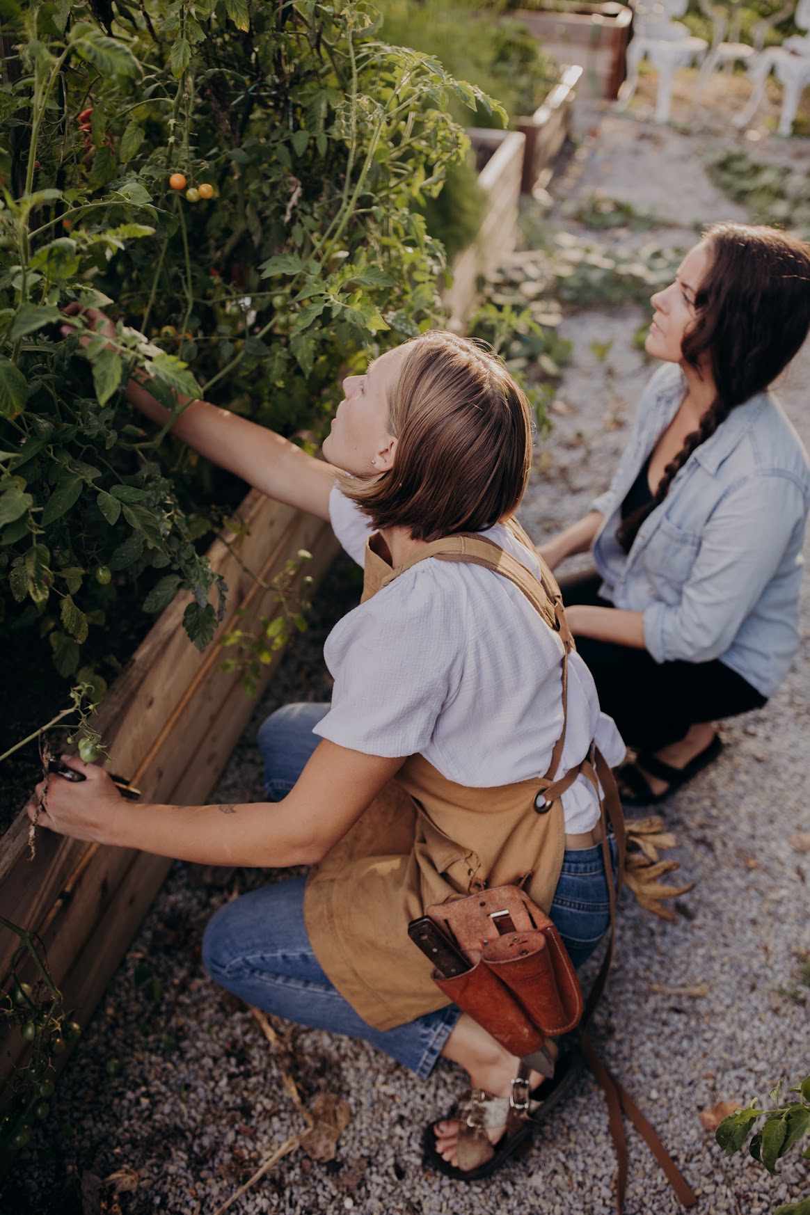 Two women are gardening outdoors, one is reaching into a plant bed, the other is kneeling nearby, both engaged in gardening.