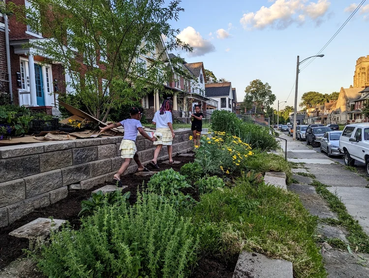 Three children walk along a sidewalk next to a garden with green plants and yellow flowers, with houses and cars in the background.