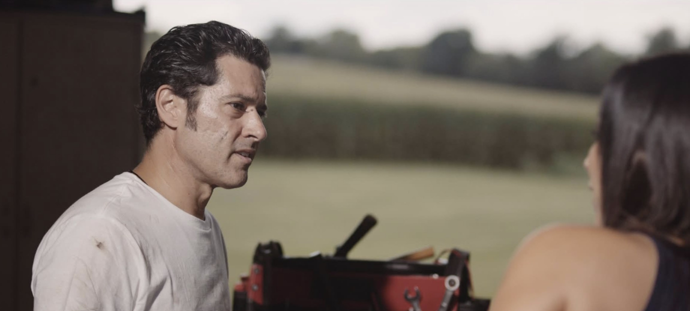 Man and woman facing each other outdoors, with a field in the background.