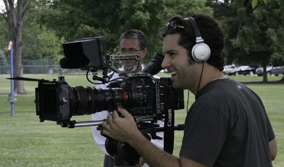 Two men operating professional video cameras outdoors in a park with trees and cars in the background.