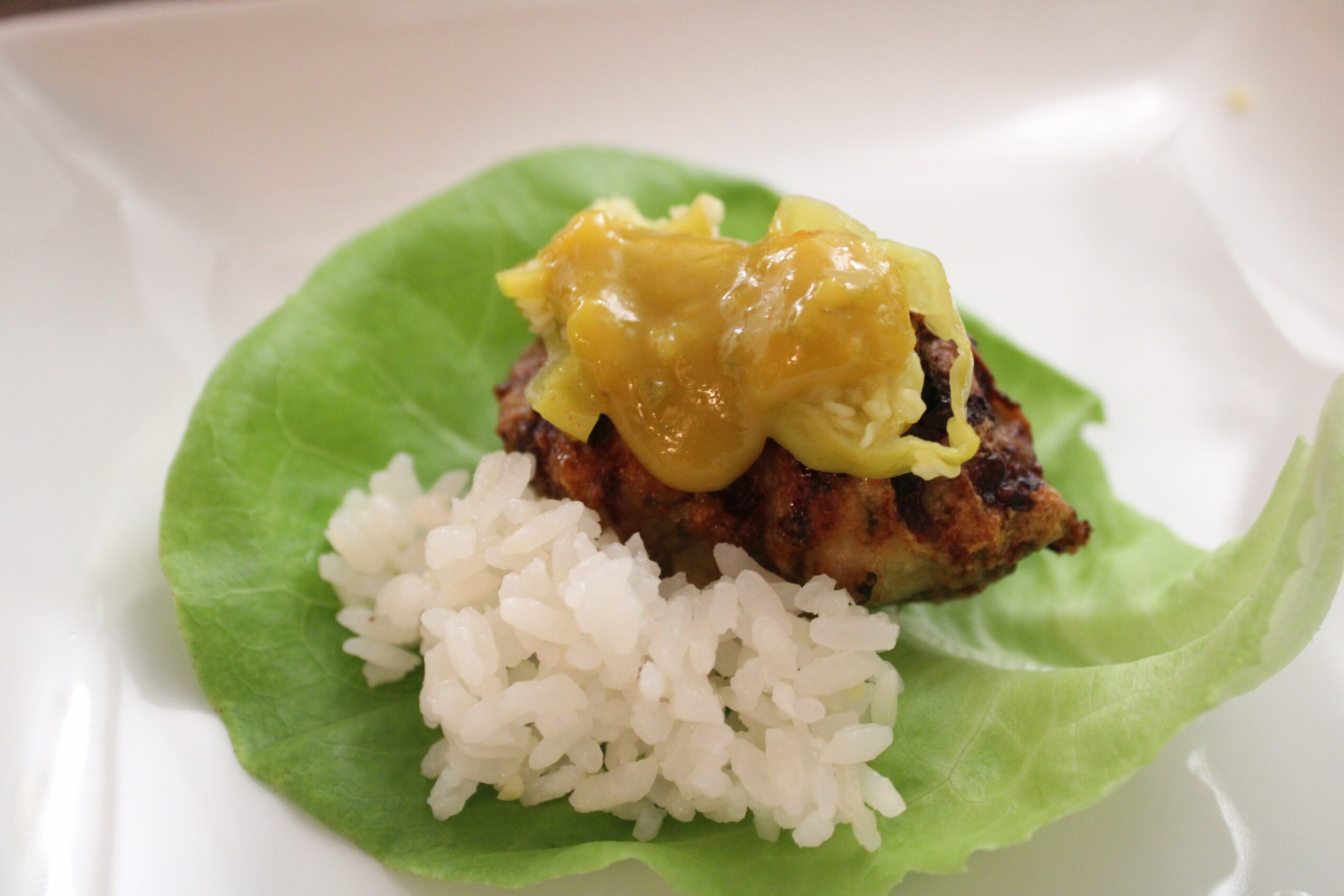 A leaf with rice, a meat patty, and yellow sauce on top, served on a white plate.