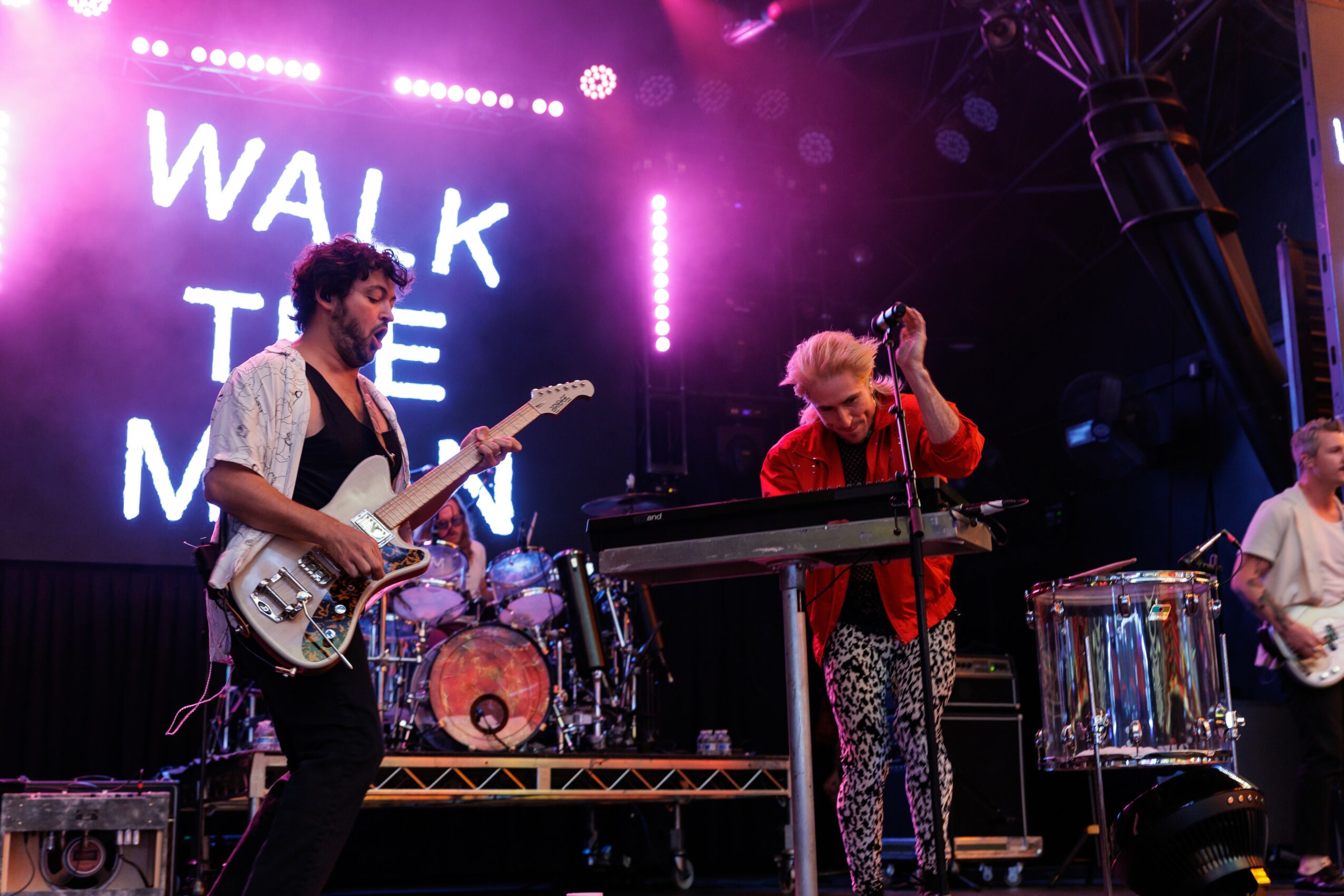 Band performing on stage with purple lighting and a sign reading 'WALK THE MOON' in the background.
