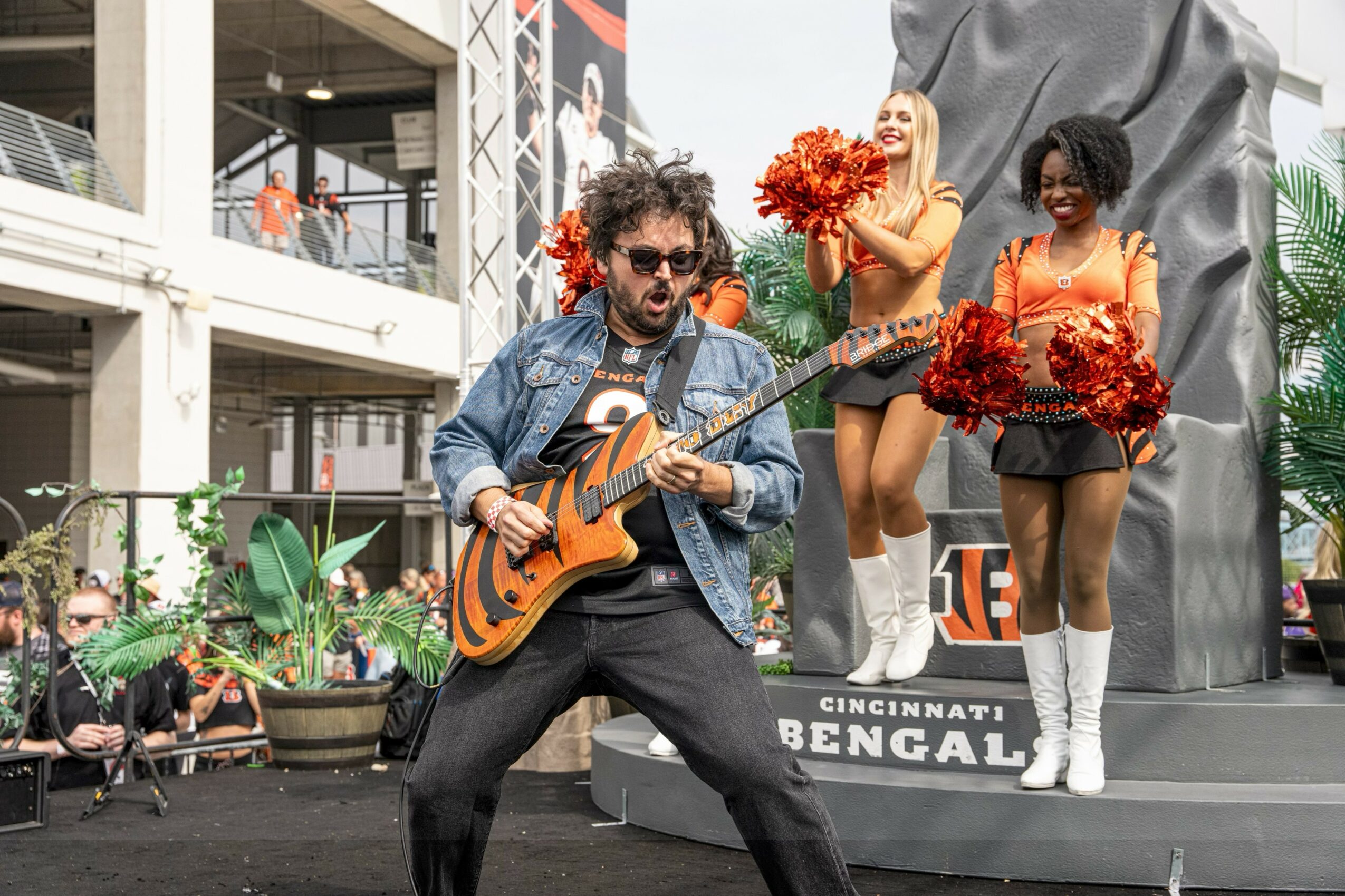 Musician playing guitar on stage with two cheerleaders holding pom-poms behind him outdoors.