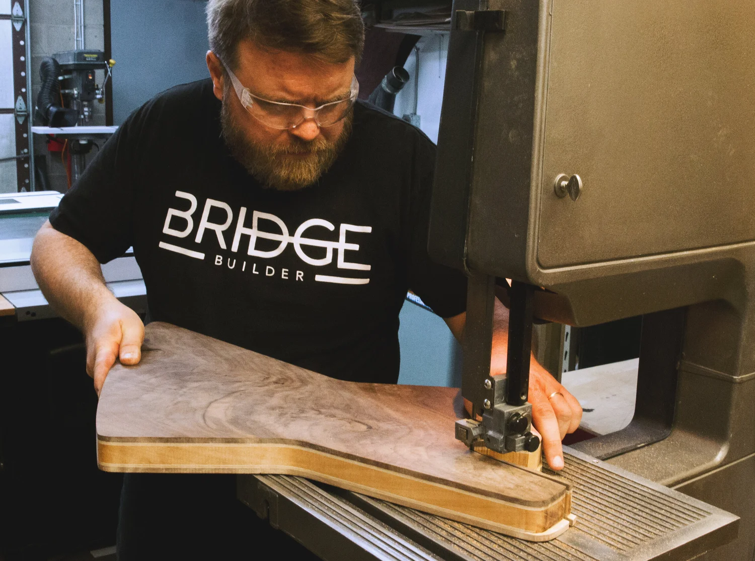 Person operating a bandsaw to cut a wooden board, wearing a black shirt with 'BRIDGE BUILDER' text.