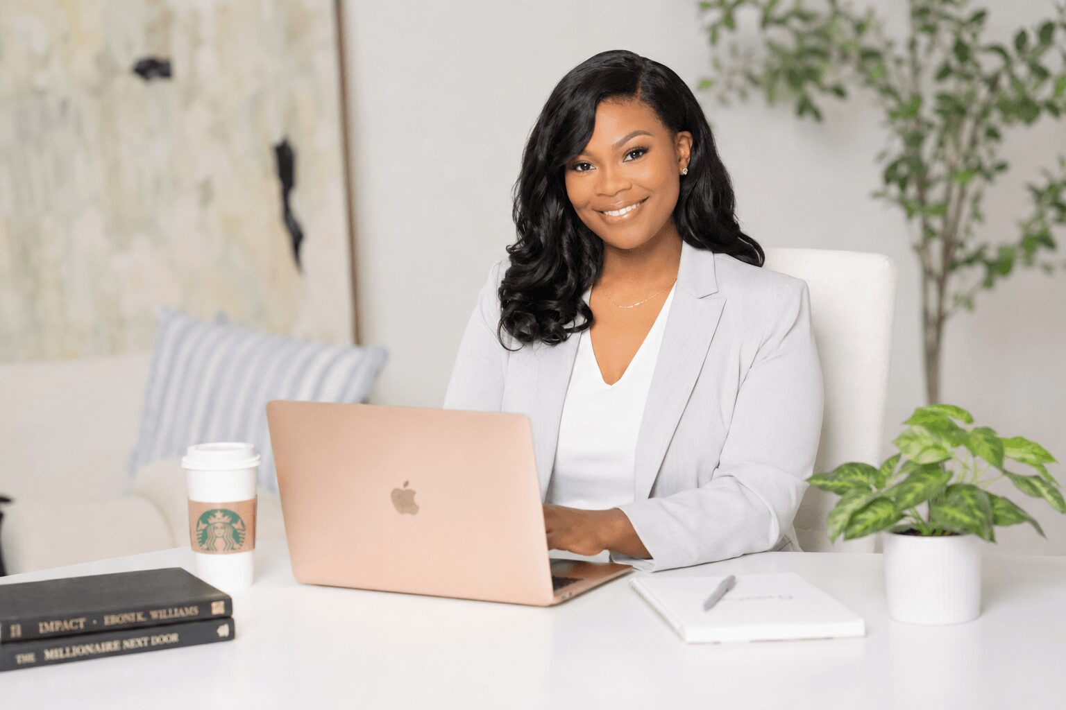Woman with dark hair in a white blazer sitting at a desk with a laptop, plant, and coffee cup, smiling.