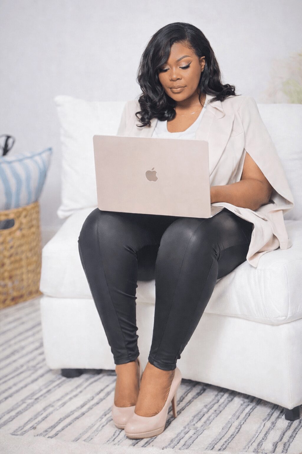 Woman sitting on a white sofa using a laptop, wearing a light-colored top and black pants, with beige high heels.