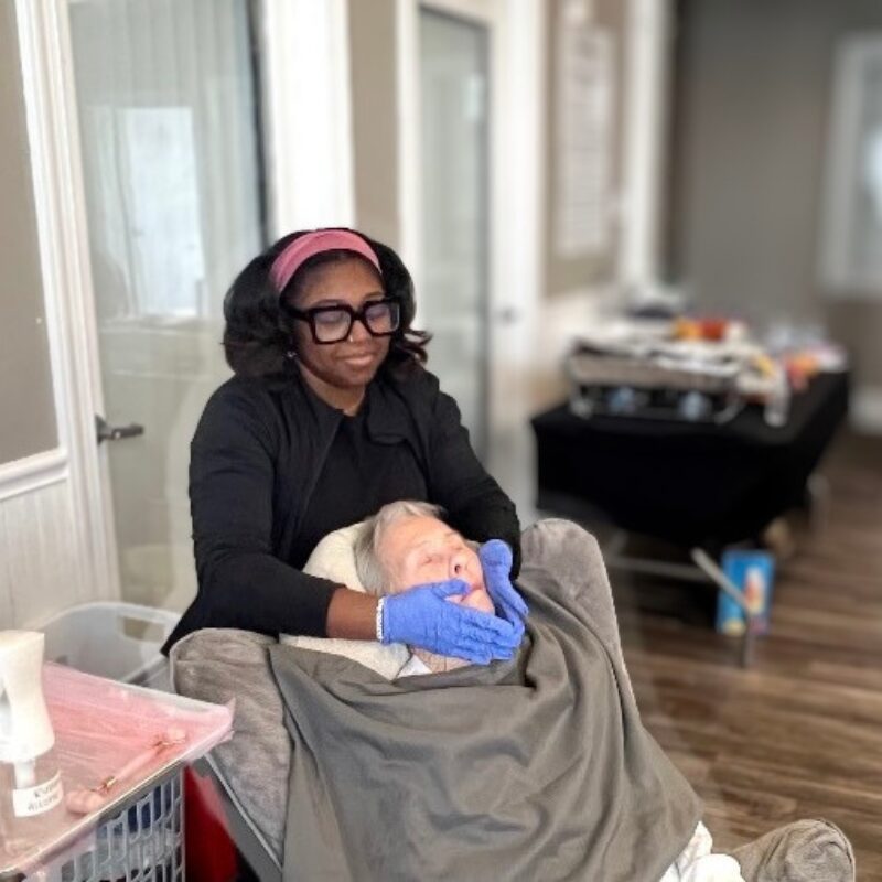 A woman wearing glasses and a pink headband holds a newborn baby wrapped in a blanket, in a room with a table and window.