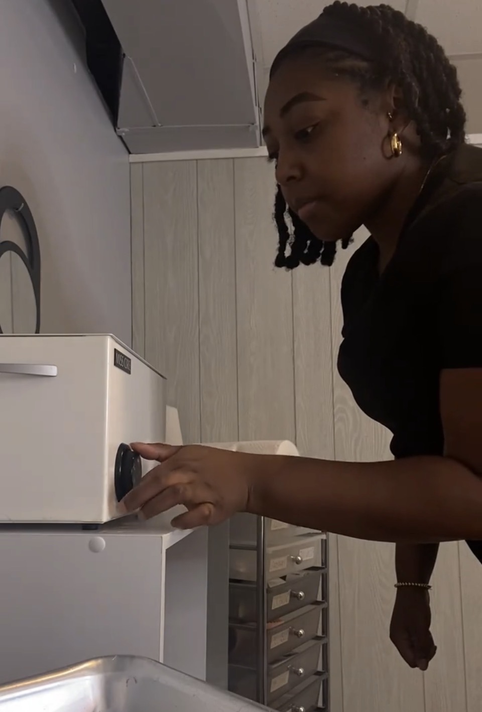 Woman adjusting a white appliance in a room with light-colored walls and a small drawer unit, side view.