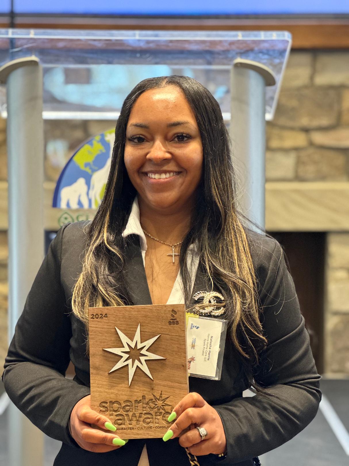 Woman with long dark hair smiling, holding an award plaque, standing indoors with a logo in the background.
