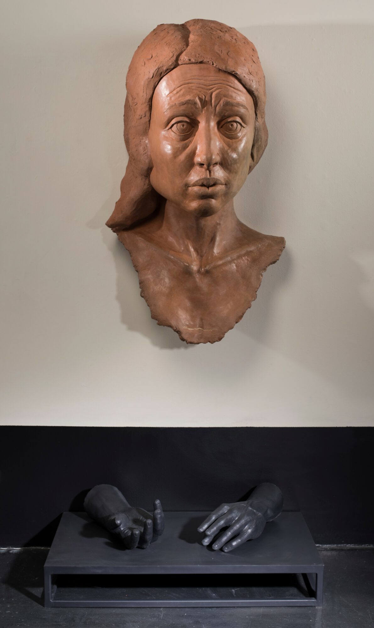 Clay bust of a woman with detailed facial features and hair, mounted on a wall, with black gloves on a table below.
