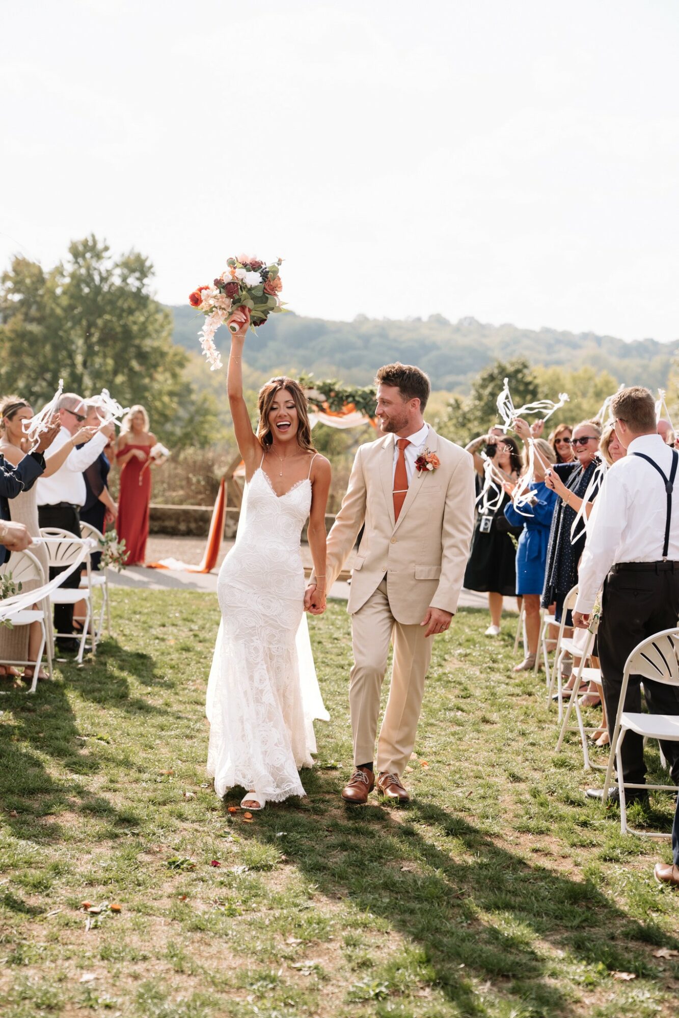 Bride and groom walking outdoors, holding hands, with guests on either side, trees and hills in background, bride holding bouquet up.