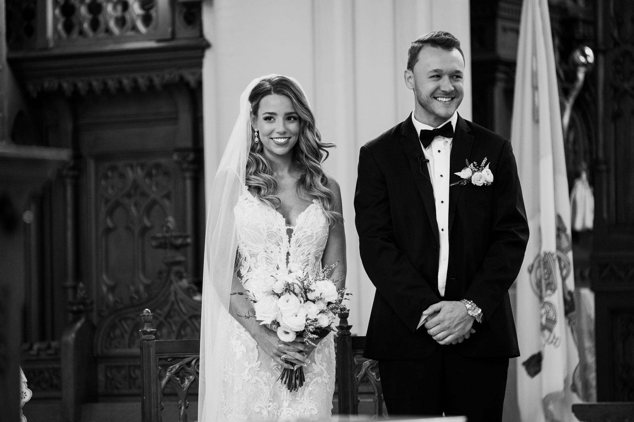 Bride and groom smiling, standing indoors with ornate woodwork and flags in background.