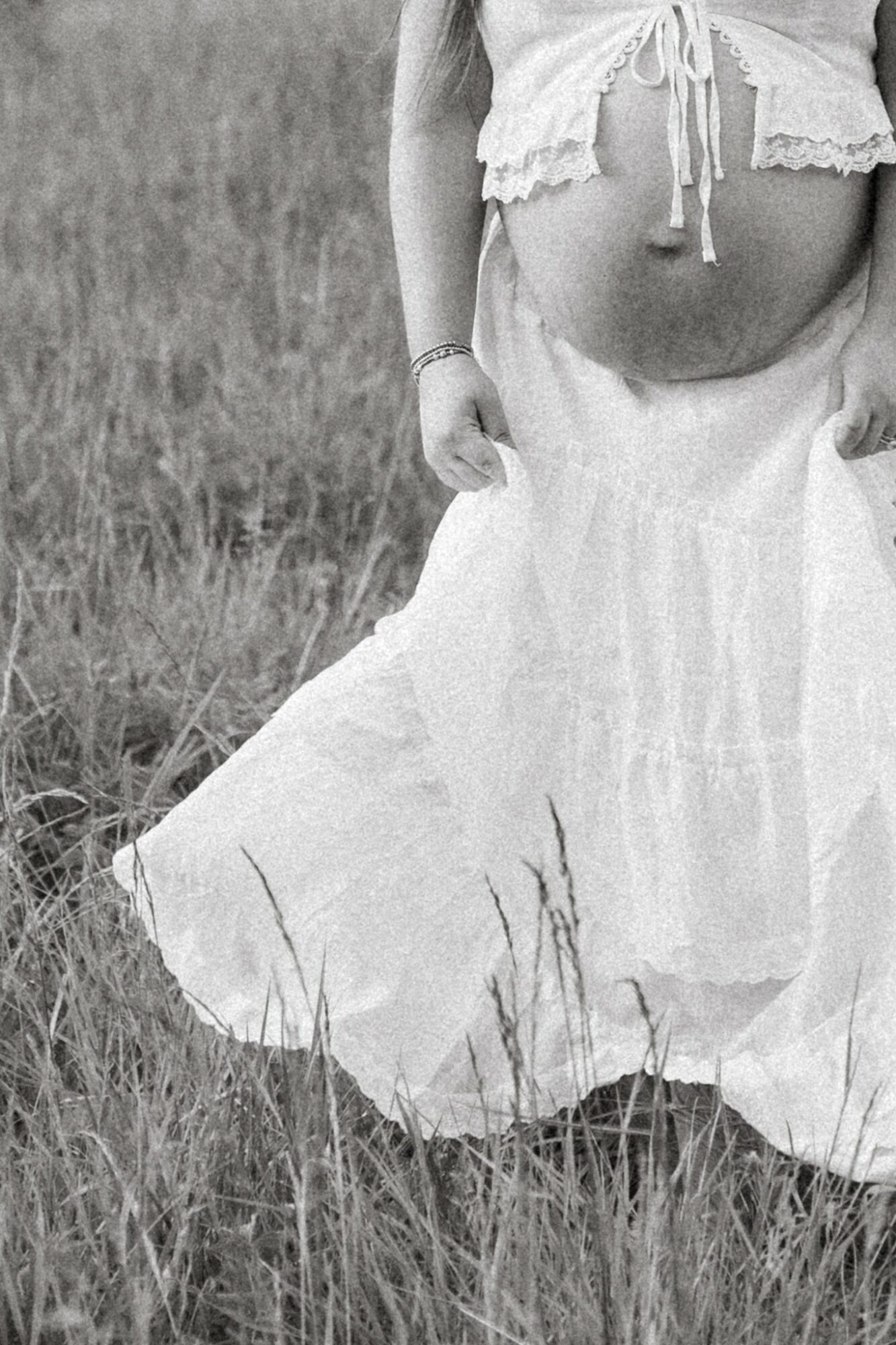 Pregnant woman standing outdoors in a grassy field, wearing a long skirt and a top tied at the waist, with her belly visible.