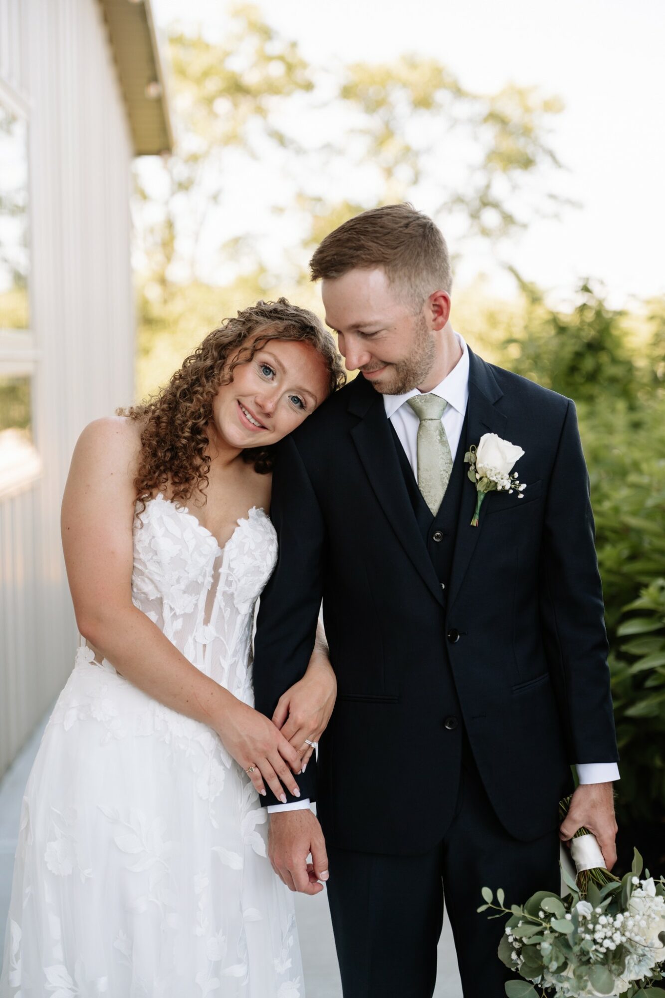 Bride and groom stand outdoors, smiling, with trees and sky in background, bride in white dress, groom in dark suit, holding flowers.