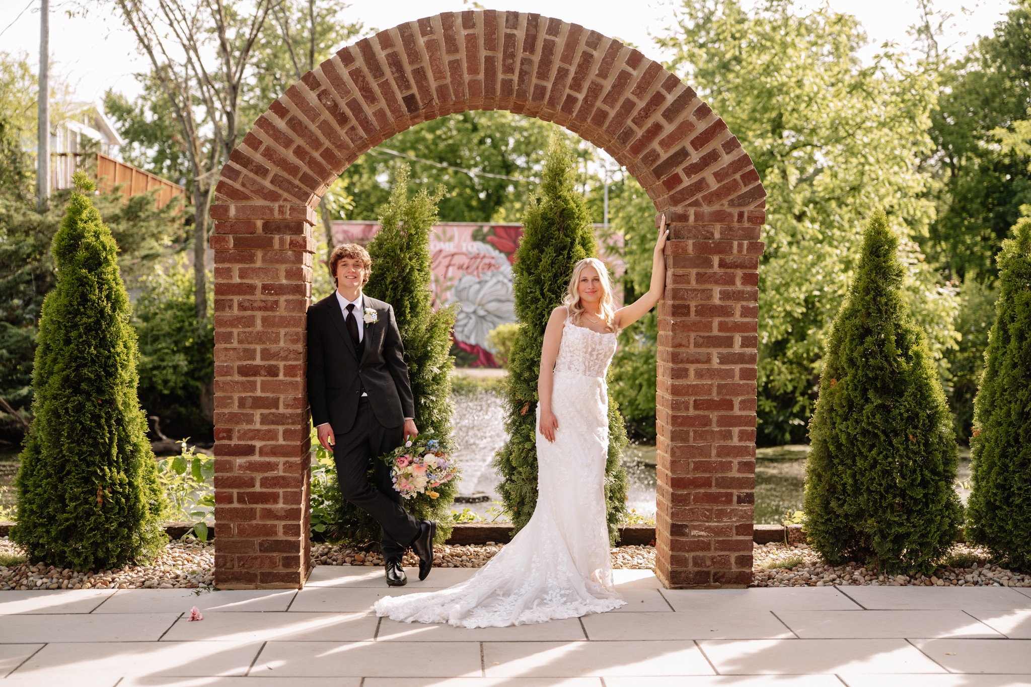 A bride and groom stand under a brick archway in a garden, surrounded by greenery and trees, on a sunny day.