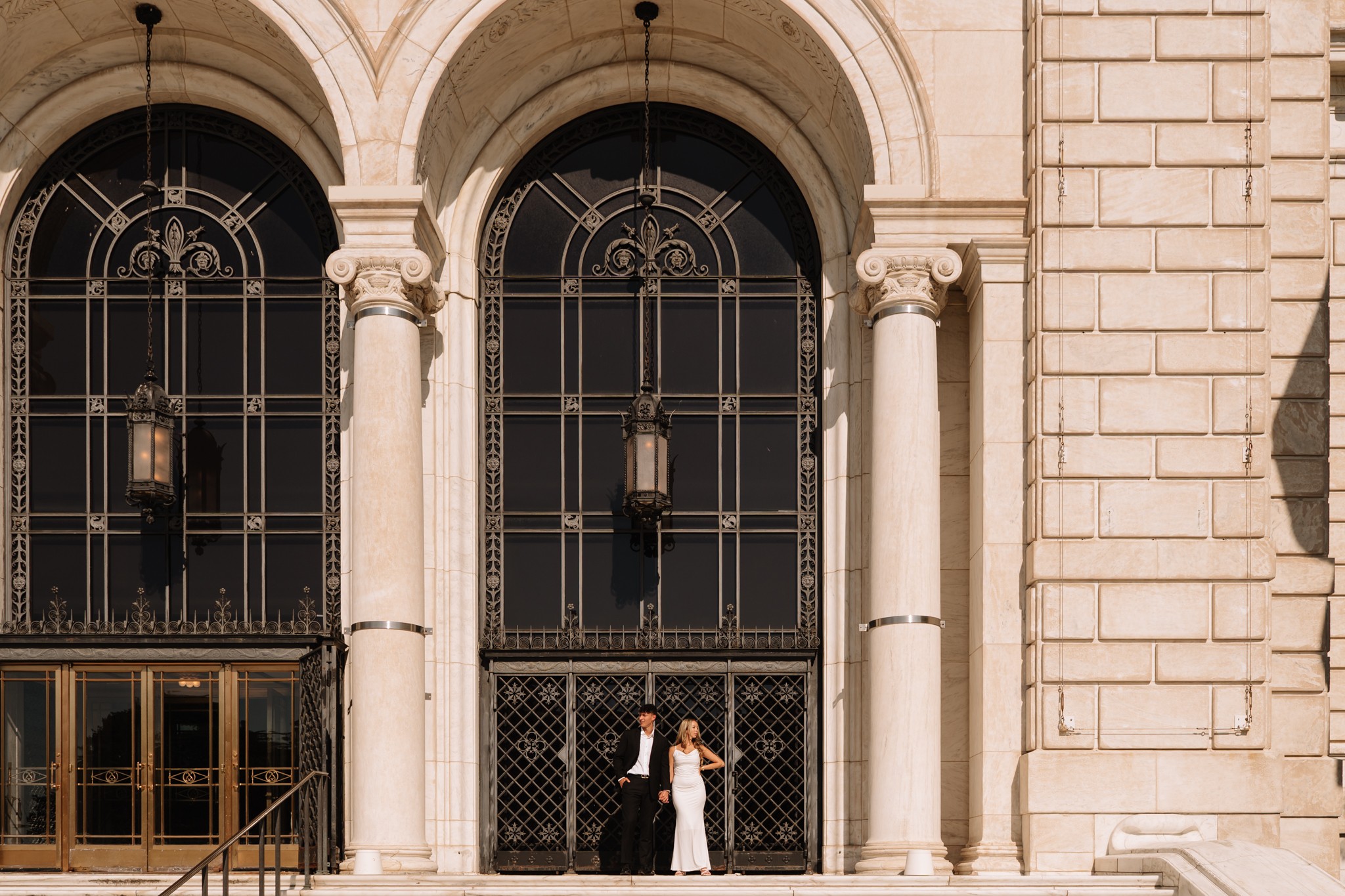 Two people stand in front of large arched windows with decorative ironwork, on a stone building facade with columns.