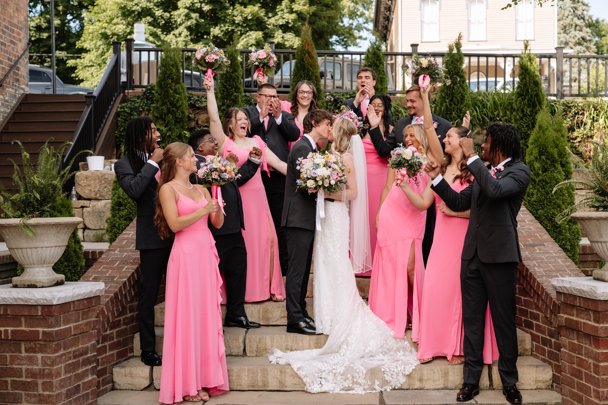 Group of people celebrating outdoors, some holding bouquets, on steps with greenery and brick walls behind.
