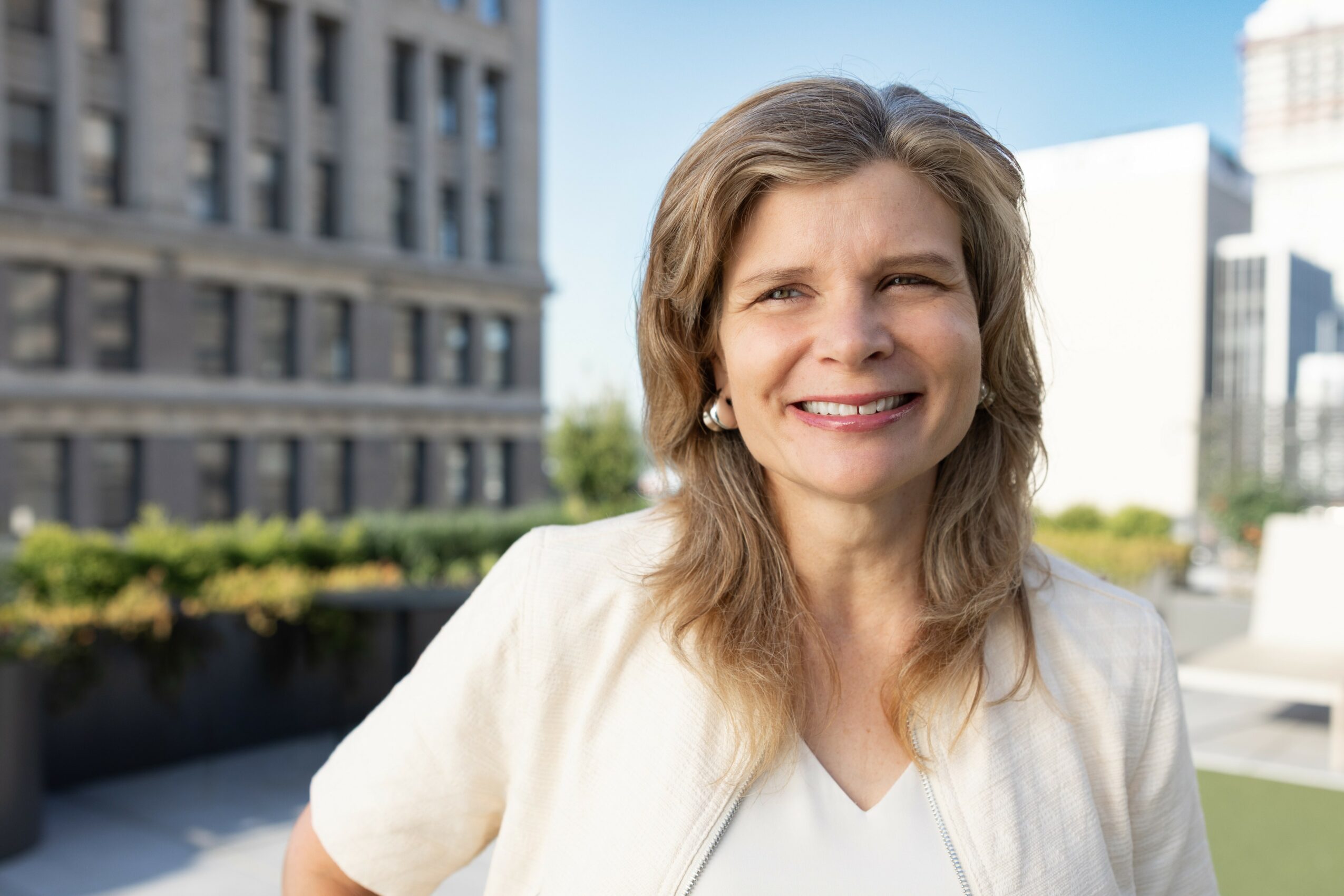 Woman with shoulder-length hair smiling outdoors in front of city buildings, wearing a light-colored top.