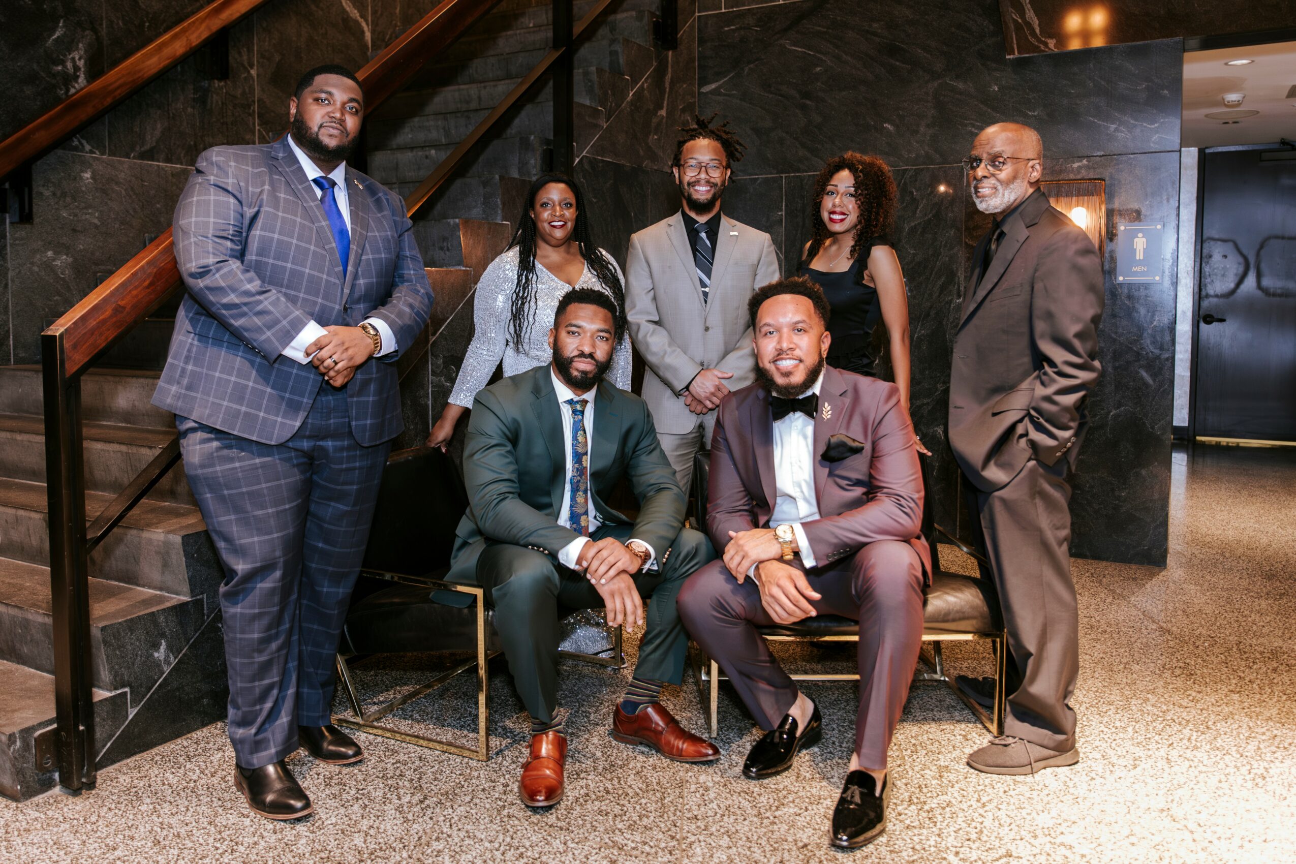 Group of seven professionally dressed people posing indoors, some standing and some seated, in front of a staircase and dark wall.
