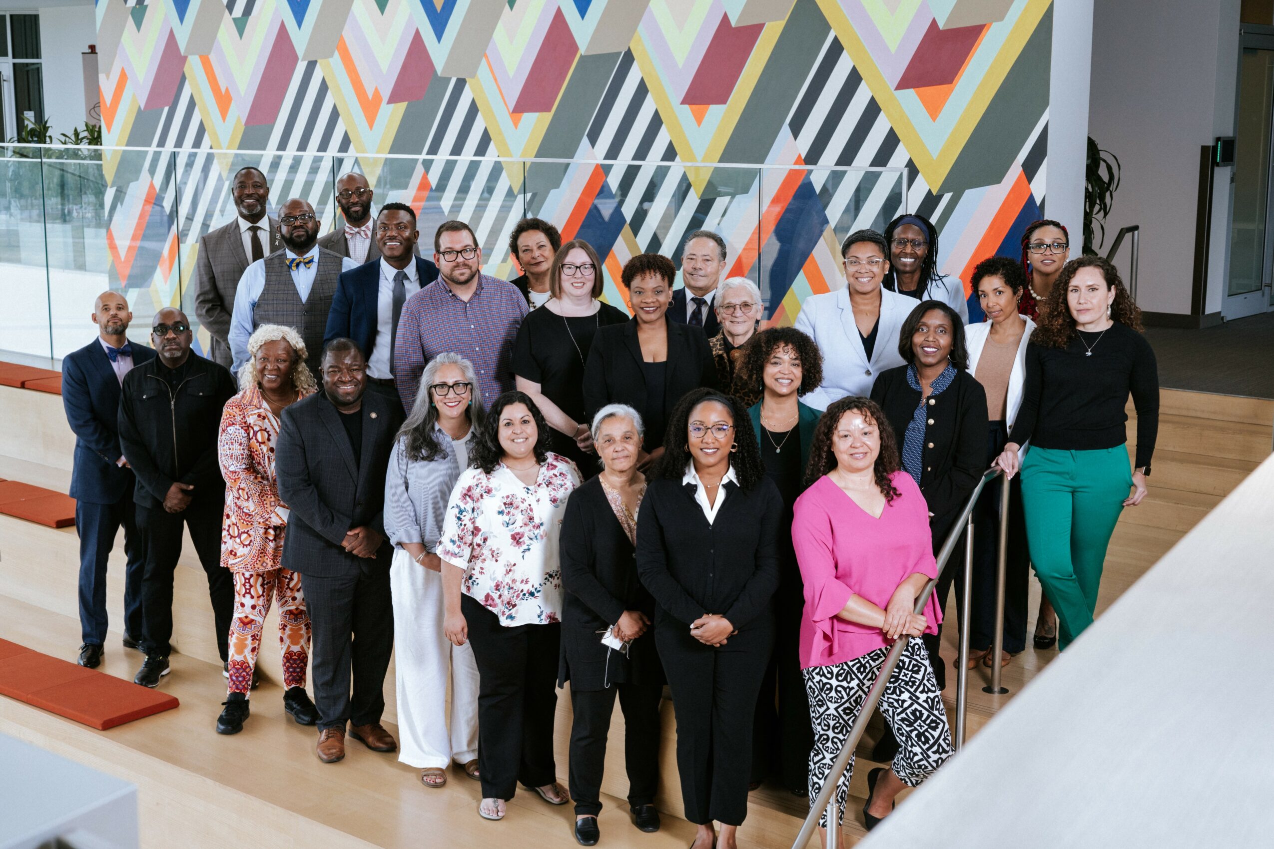 Group of diverse people standing together indoors in front of a colorful geometric wall, smiling for a photo.