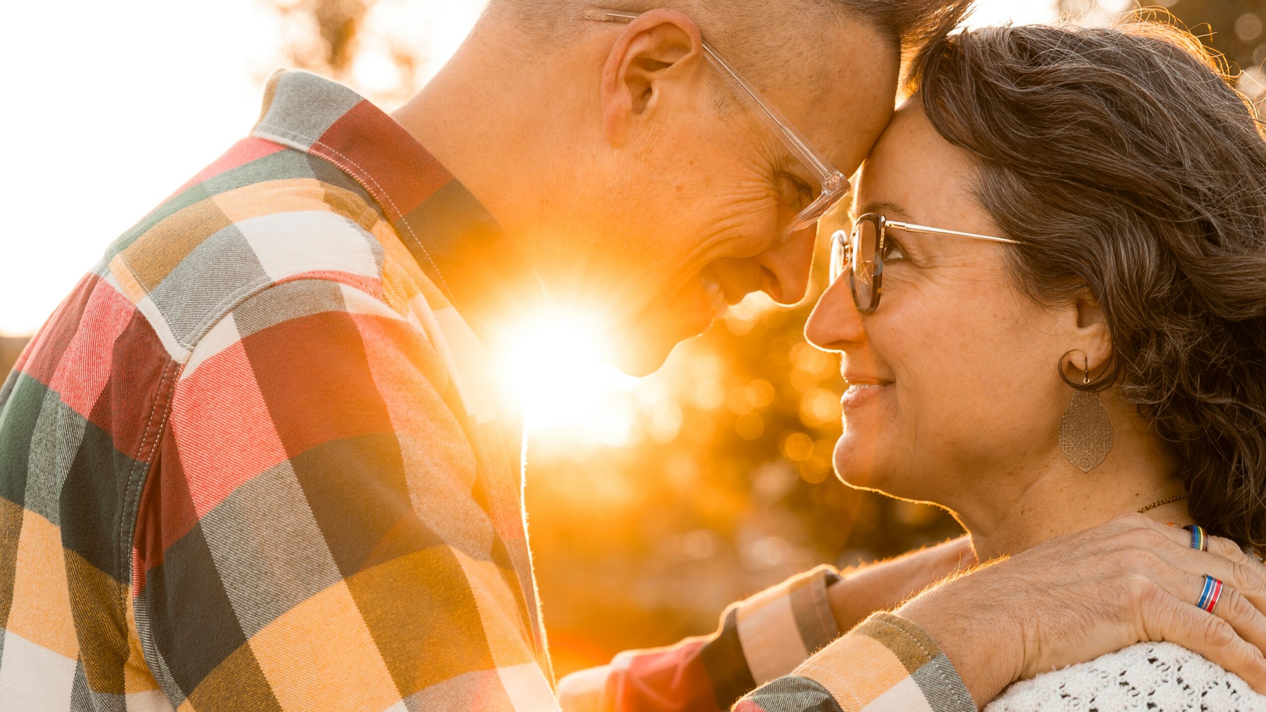 A couple faces close together outdoors during sunset, smiling and touching foreheads, with sunlight behind them.