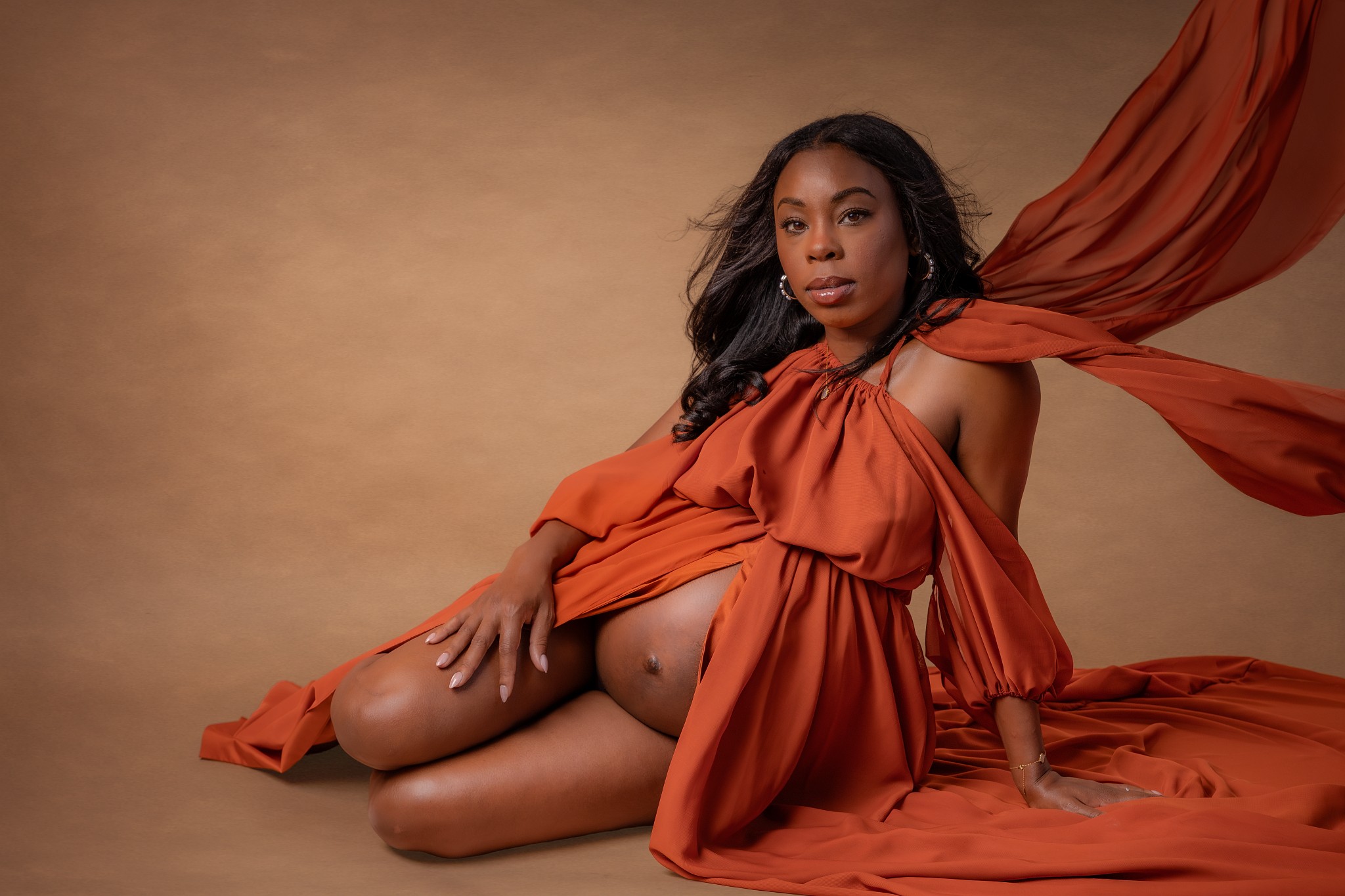 Woman with long dark hair in an orange dress, sitting on the floor with a beige background.