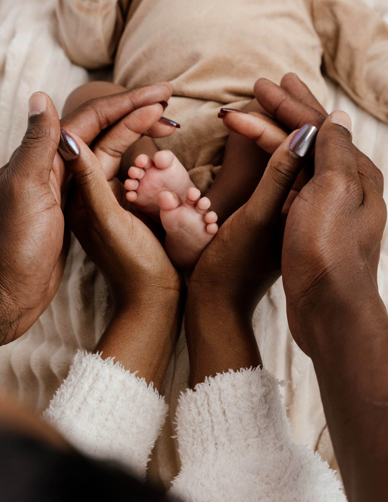 Two adult hands gently hold a tiny baby's feet, with a baby's torso visible in the background, all on a soft surface.