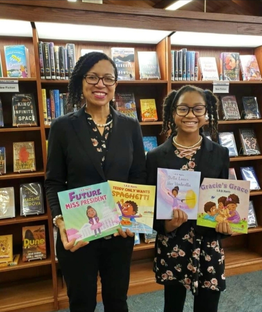 Two women holding children's books in a library, smiling, with bookshelves in the background.