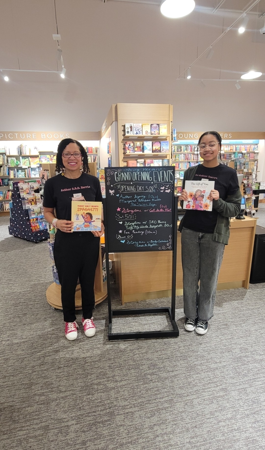 Two women stand on either side of a blackboard in a bookstore, holding books and smiling.
