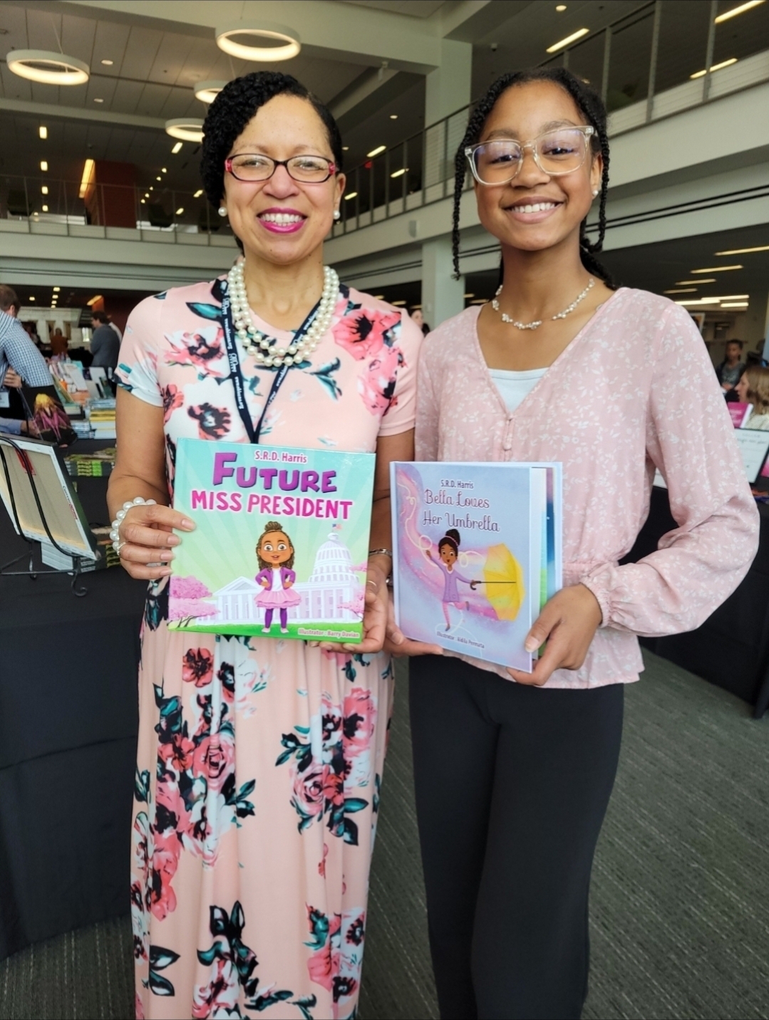 Two women smiling and holding children's books in an indoor setting with multiple levels and people in the background.