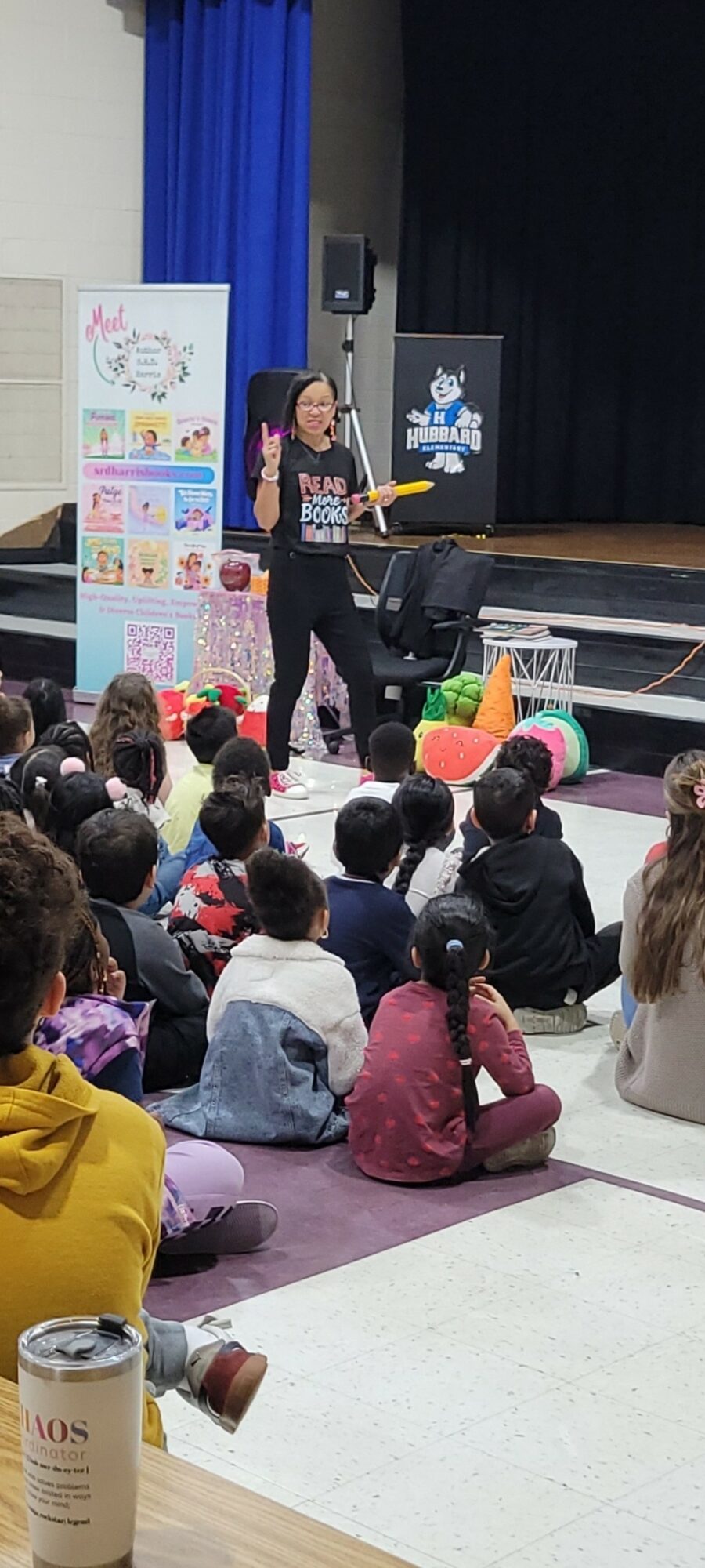Woman speaking to children seated on the floor in a room with stage, posters, and blue curtains.